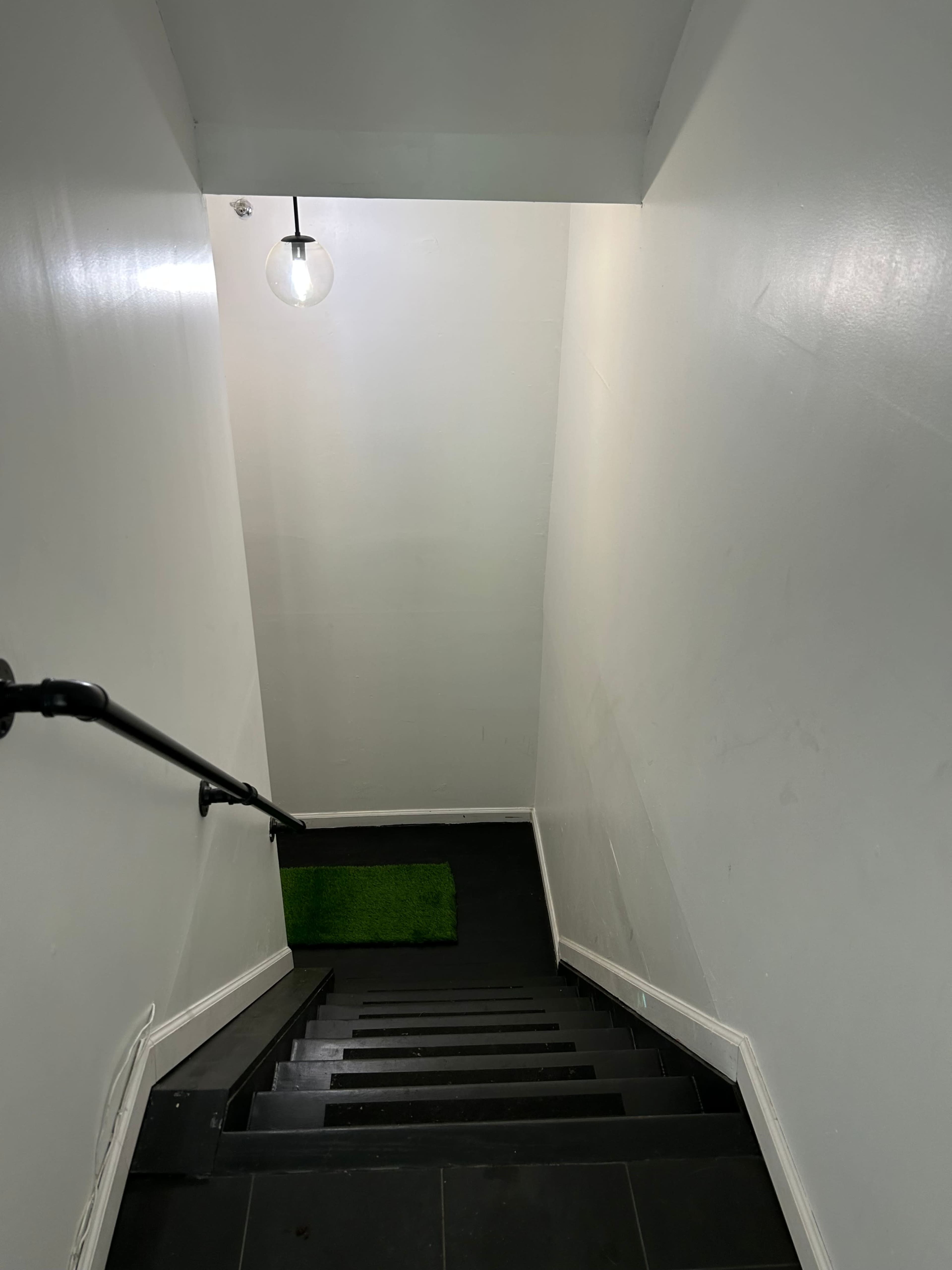A staircase descends into a hallway with a green mat at the bottom and a pendant light hanging above.
