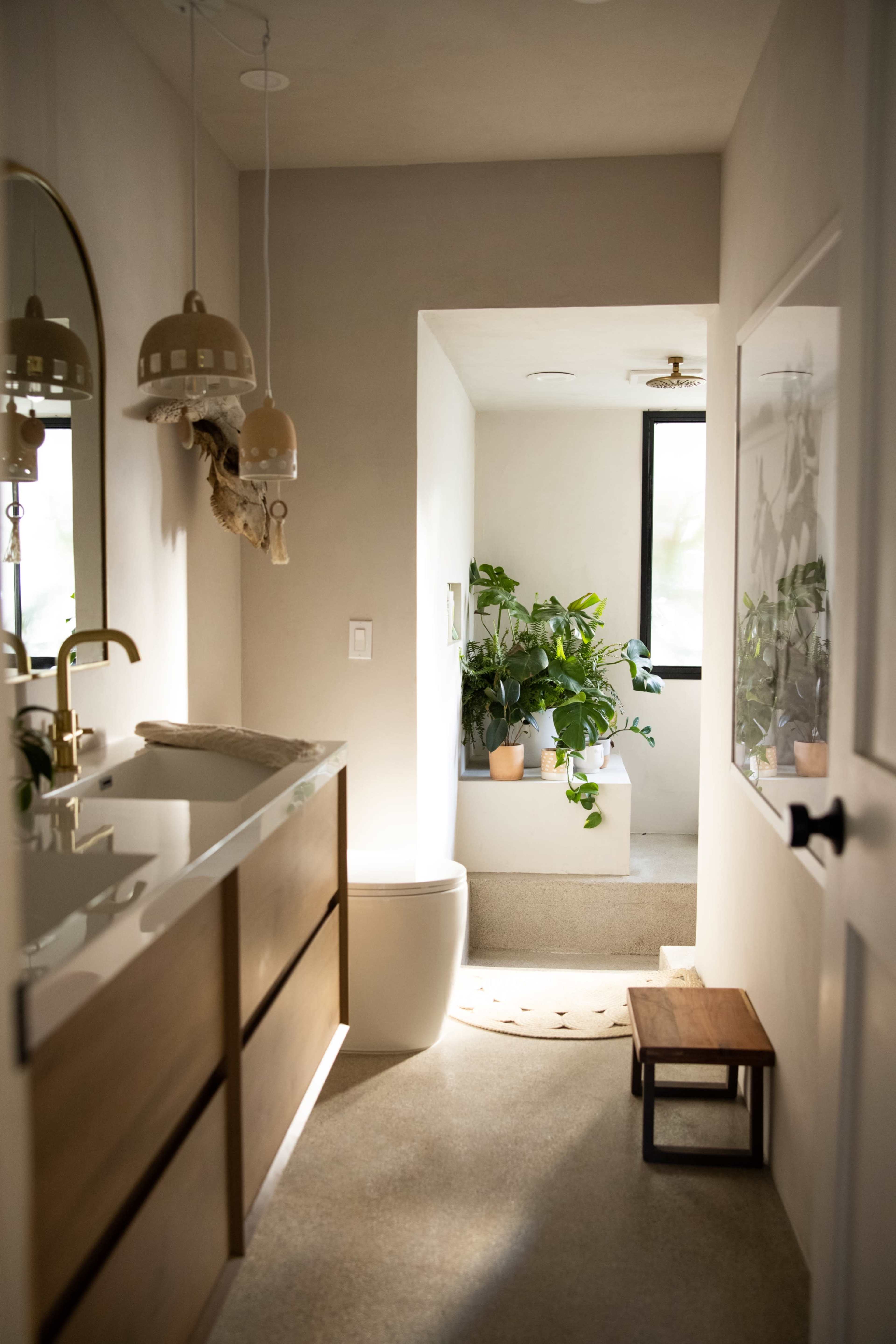 A streamlined bathroom features a wooden vanity, a modern toilet, and a sunlit tub area with potted plants.