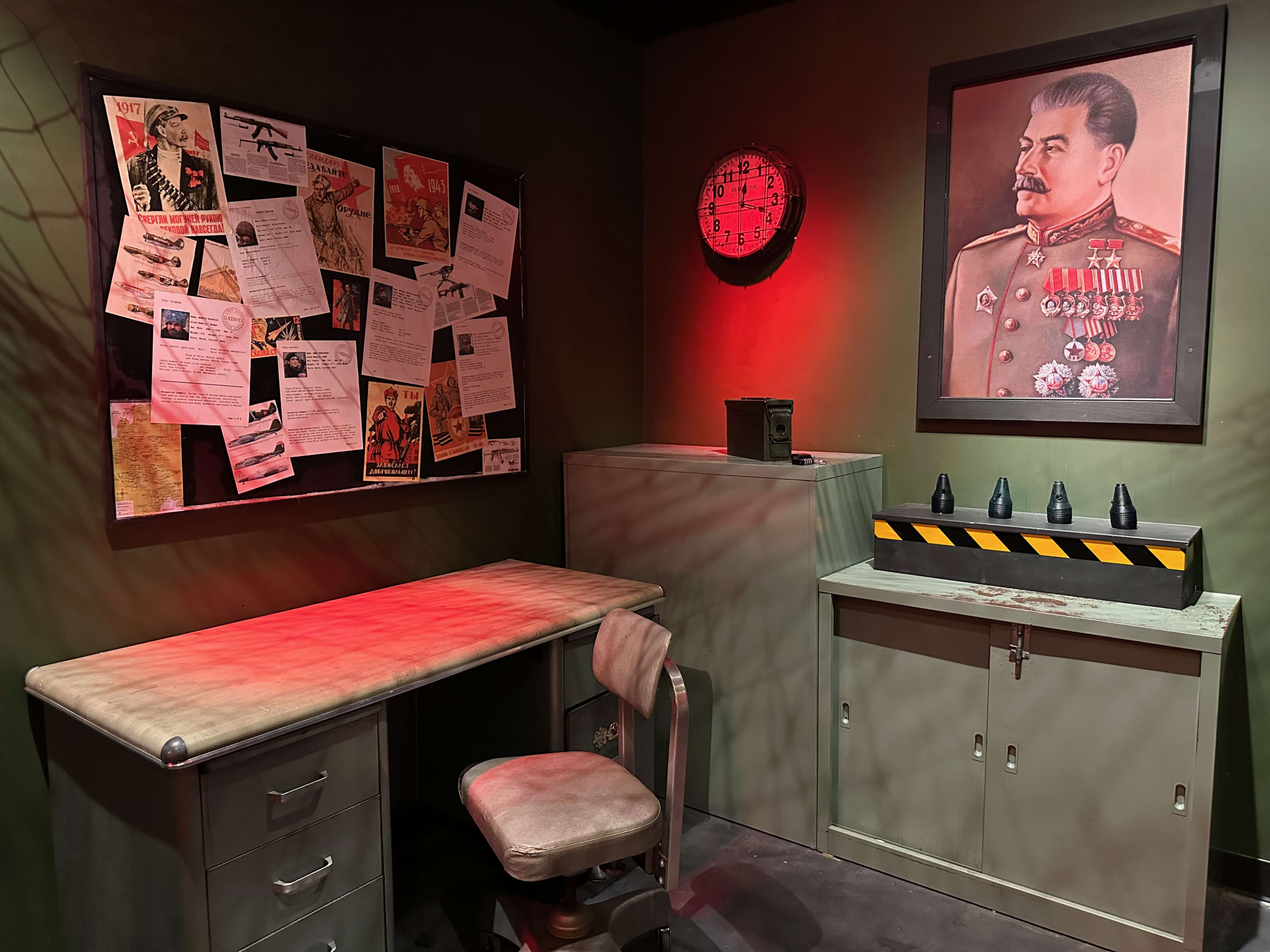 The image shows a dimly lit historical room featuring a desk and chair, a bulletin board filled with documents and images, a clock on the wall, and a portrait of a military figure, along with a cabinet and a device with warning stripes.