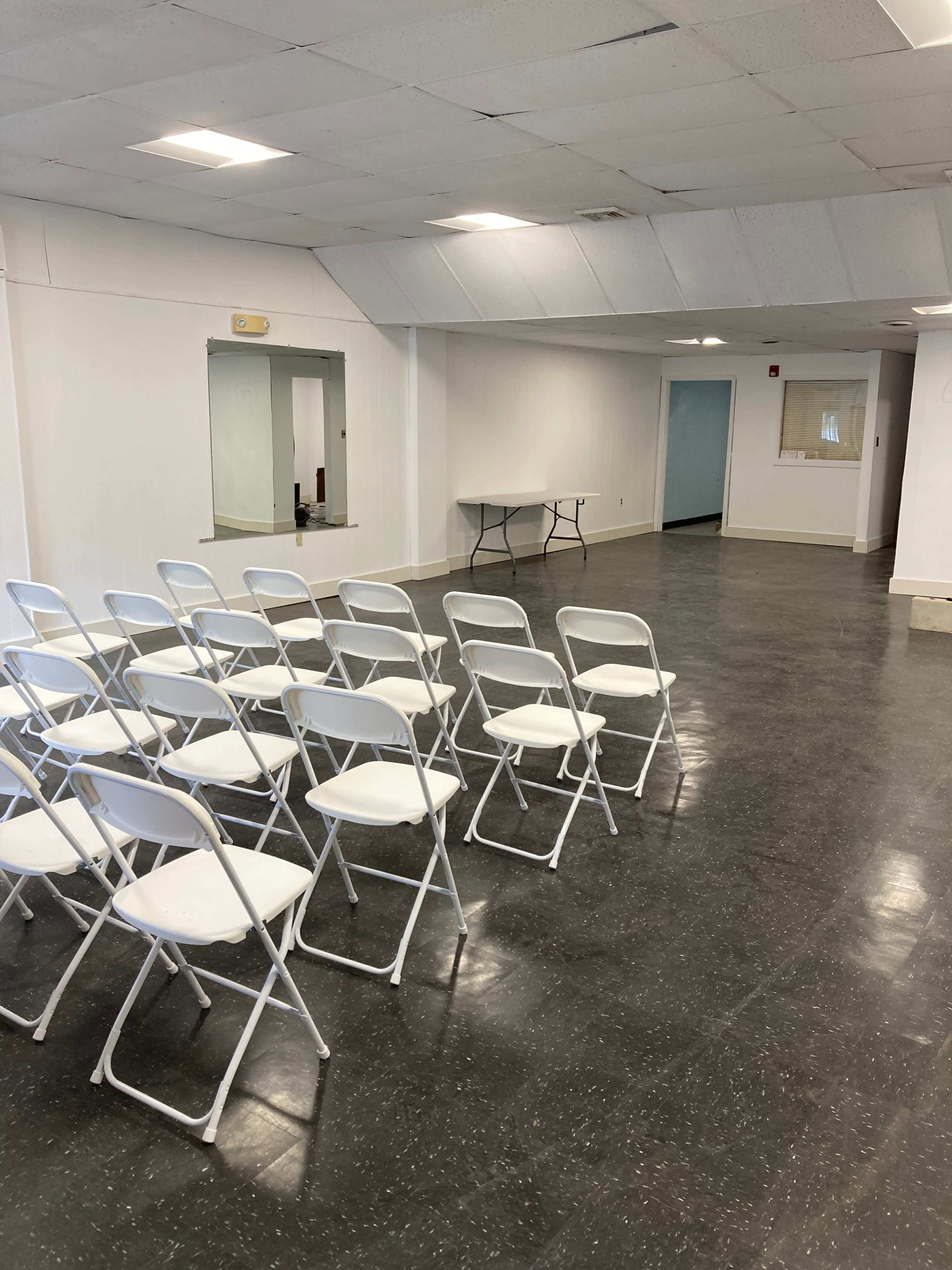 An empty room with a row of white folding chairs facing a table against a plain wall.