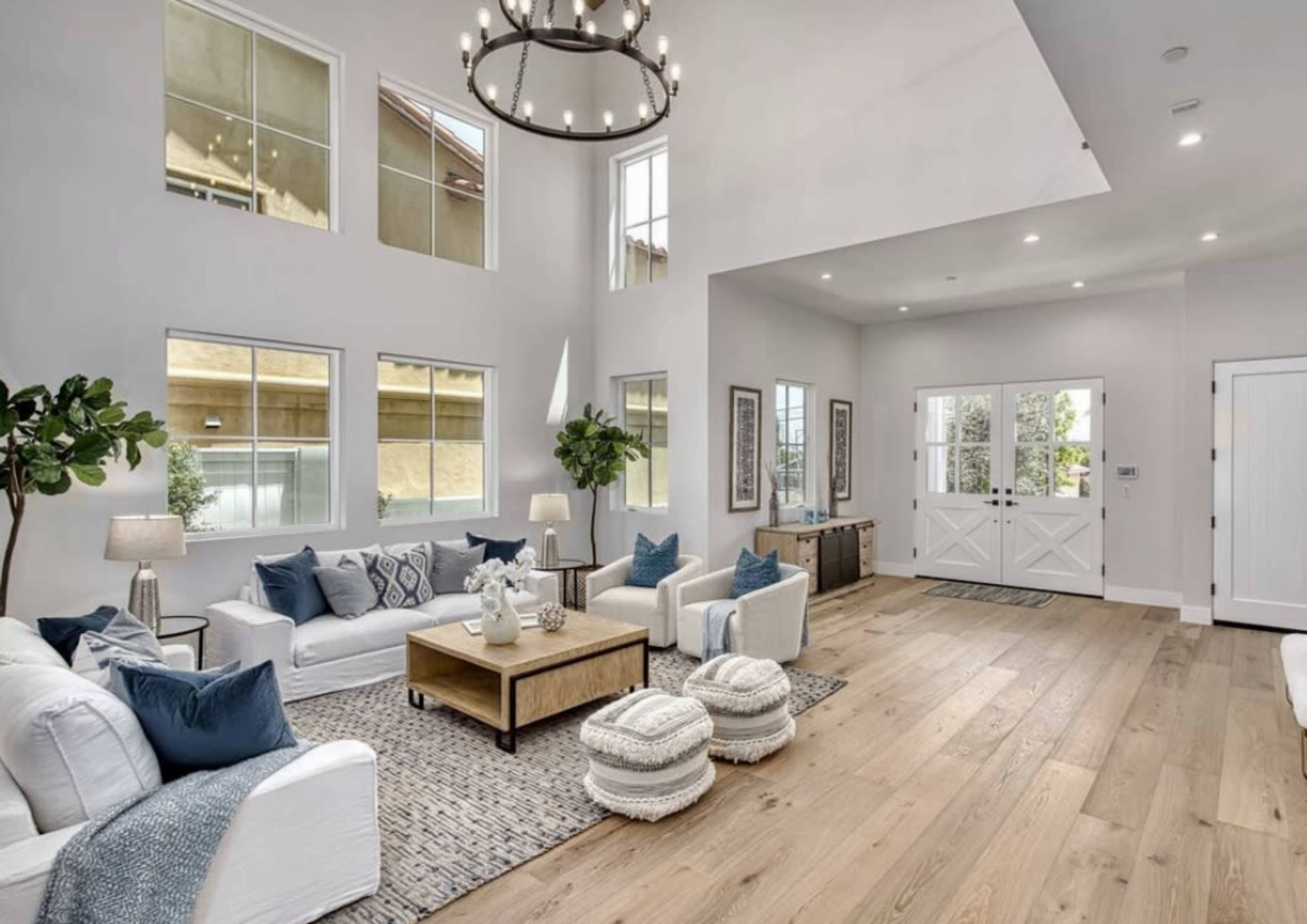 A bright living room features white furniture, large windows, a wooden floor, and a welcoming entrance with double doors.
