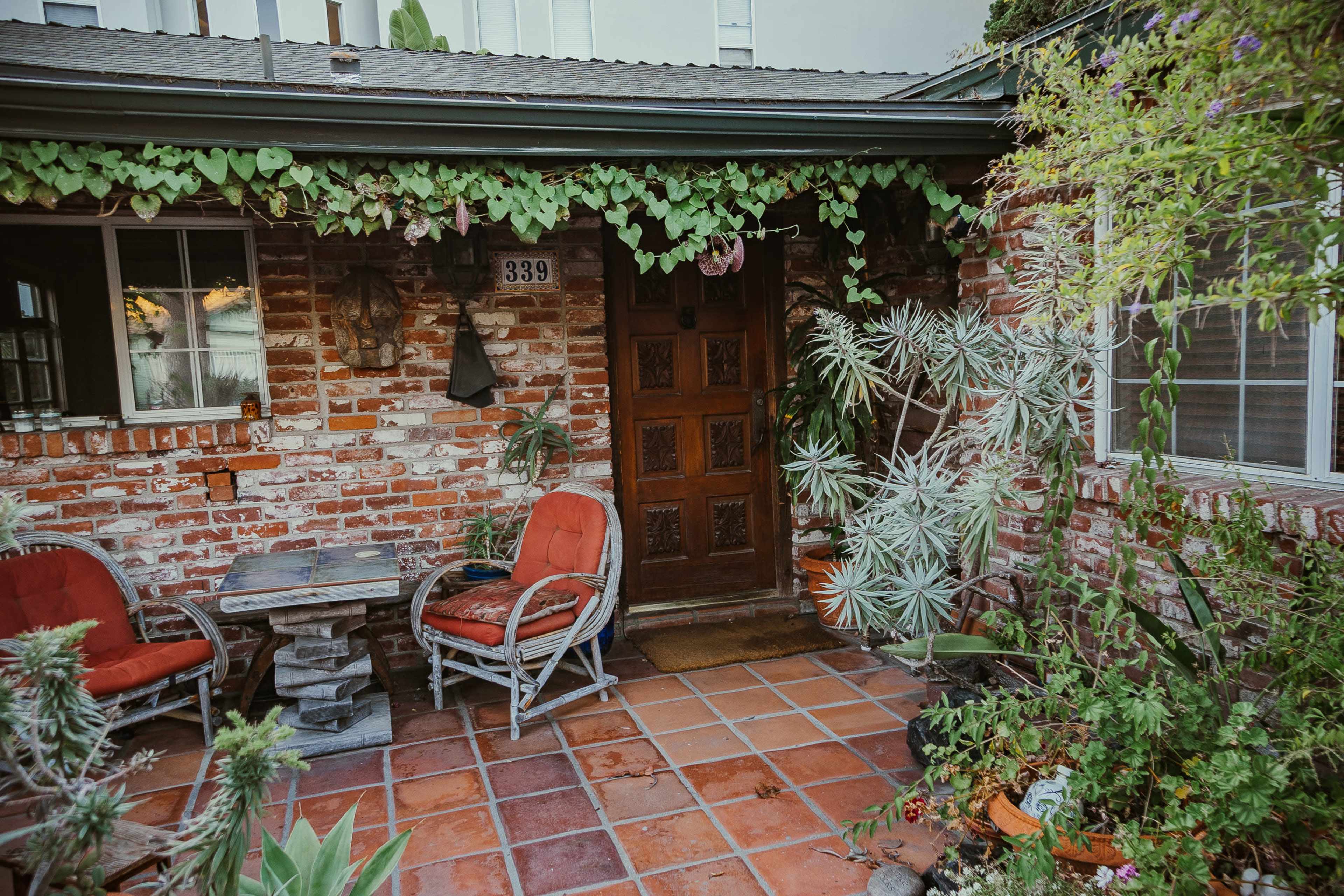 A brick patio with two red chairs, a small table, and various plants surrounding a wooden door adorned with vines.