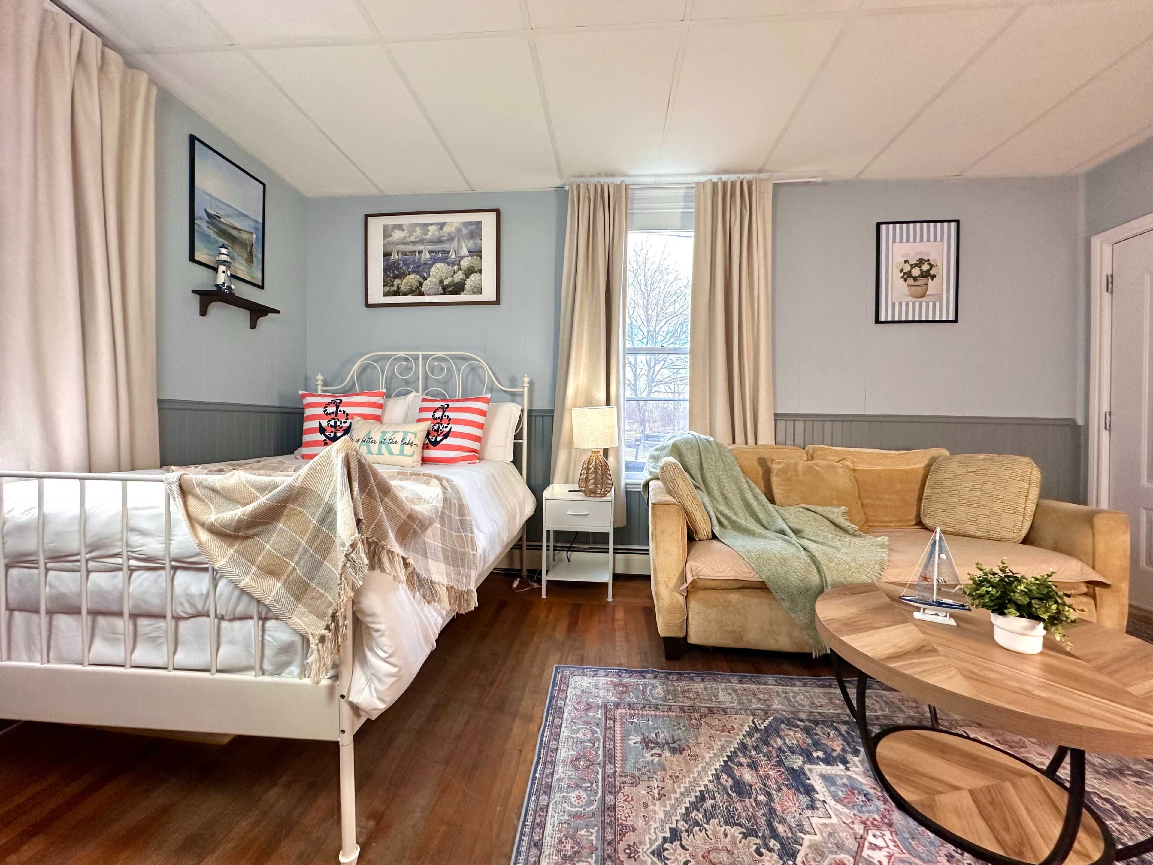 The image shows a cozy bedroom with a bed, a sofa, a coffee table, and large windows covered with curtains.