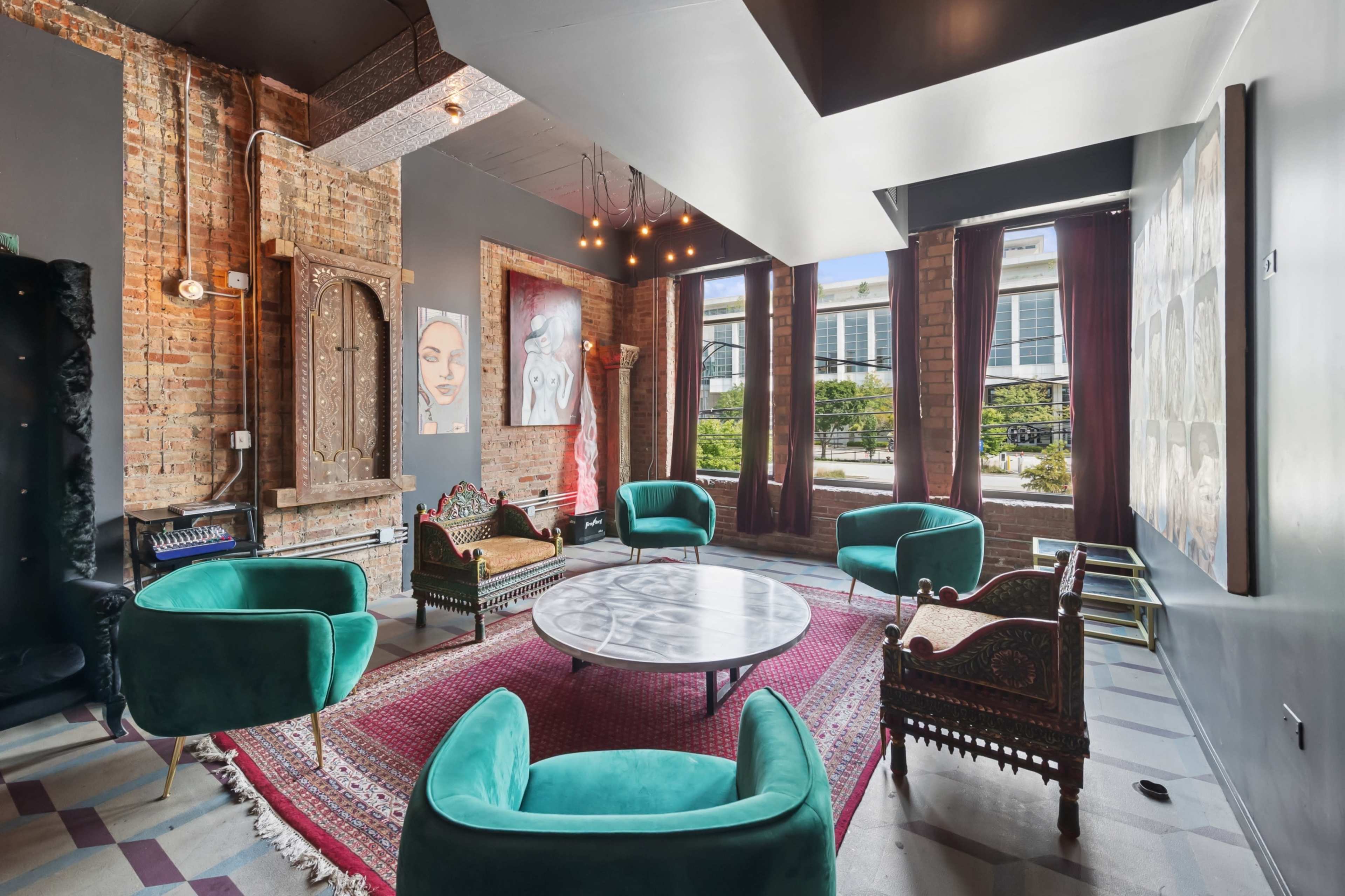The image depicts a stylish lounge area featuring four green chairs and a round coffee table, surrounded by exposed brick walls and large windows.