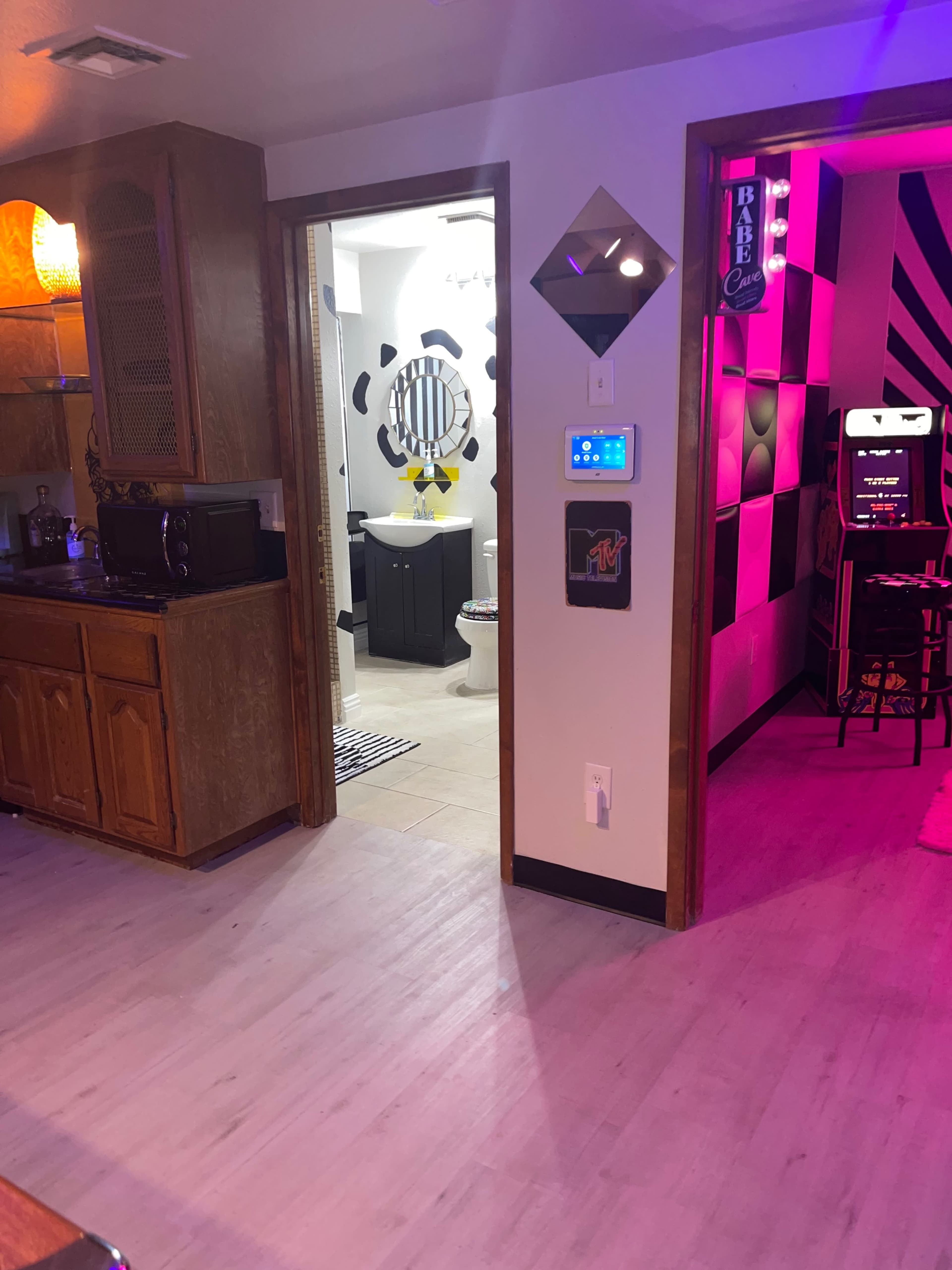 A view from a kitchen area into a hallway leading to a bathroom on the left and a room with colorful lighting on the right.