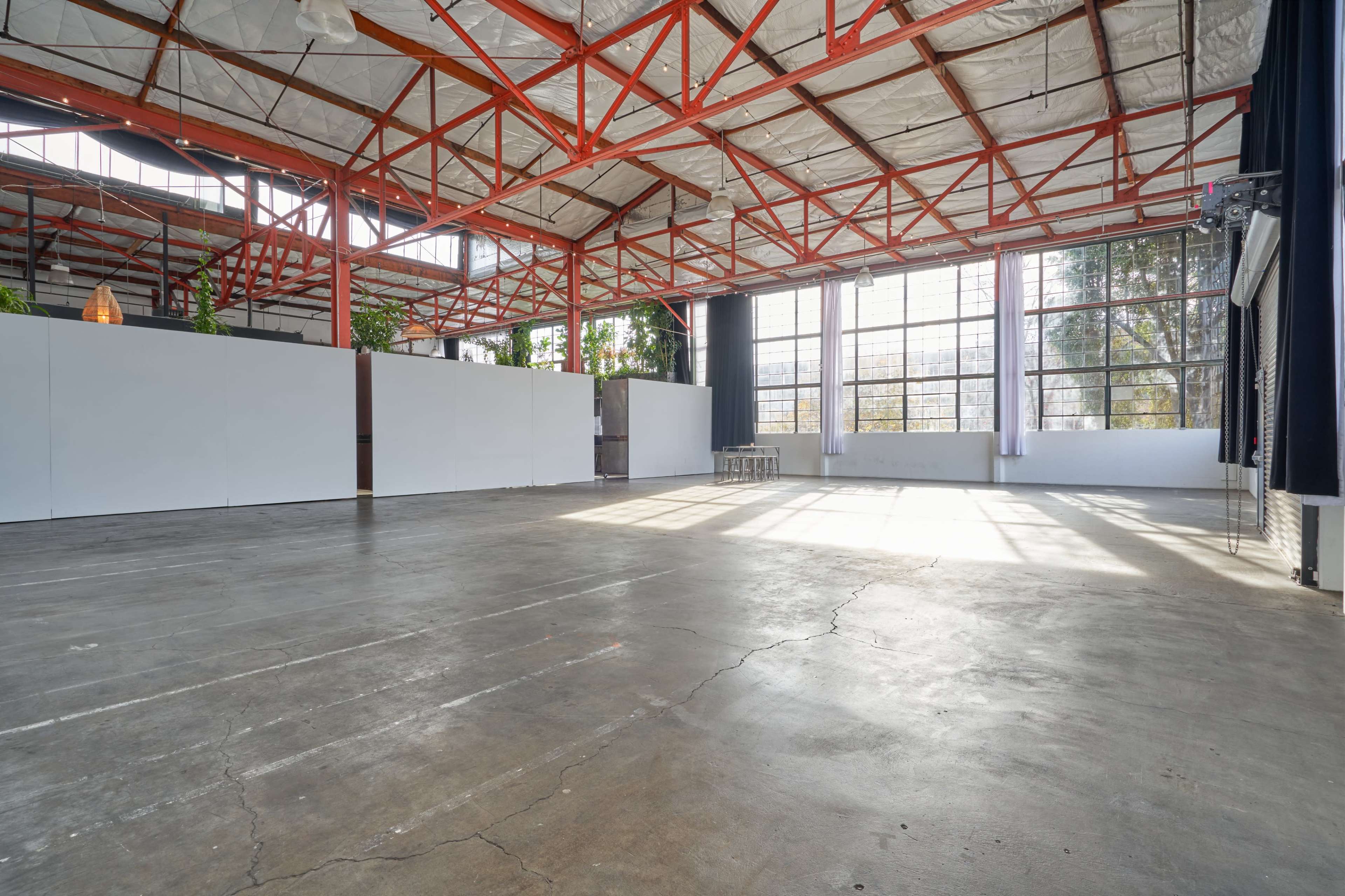 A spacious industrial venue with high ceilings, large windows, and a concrete floor, featuring white wall partitions and greenery.