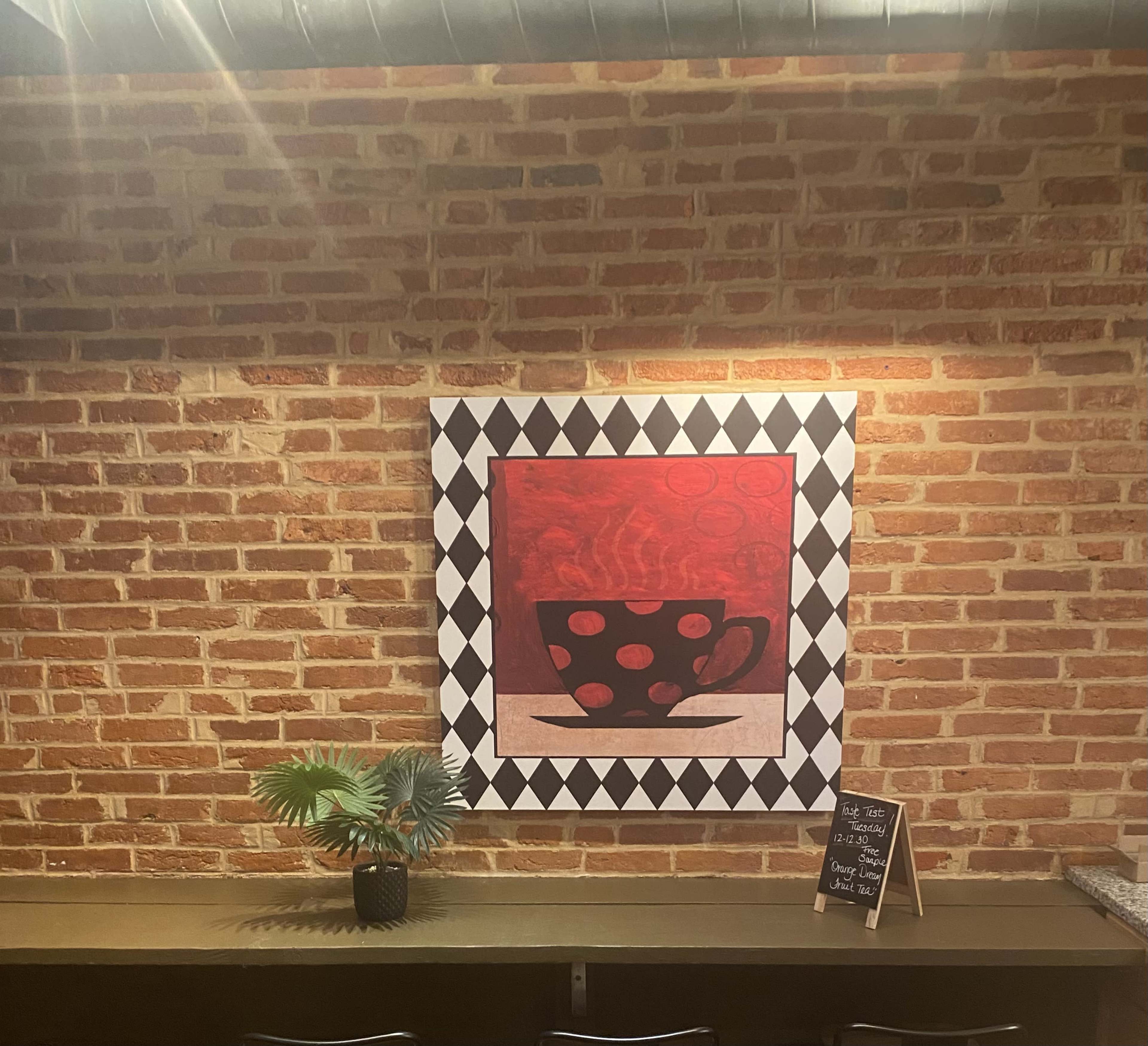 The image features a brick wall with a large, framed artwork of a polka-dotted cup on a red background, accompanied by a small potted plant and a chalkboard sign on a countertop below.