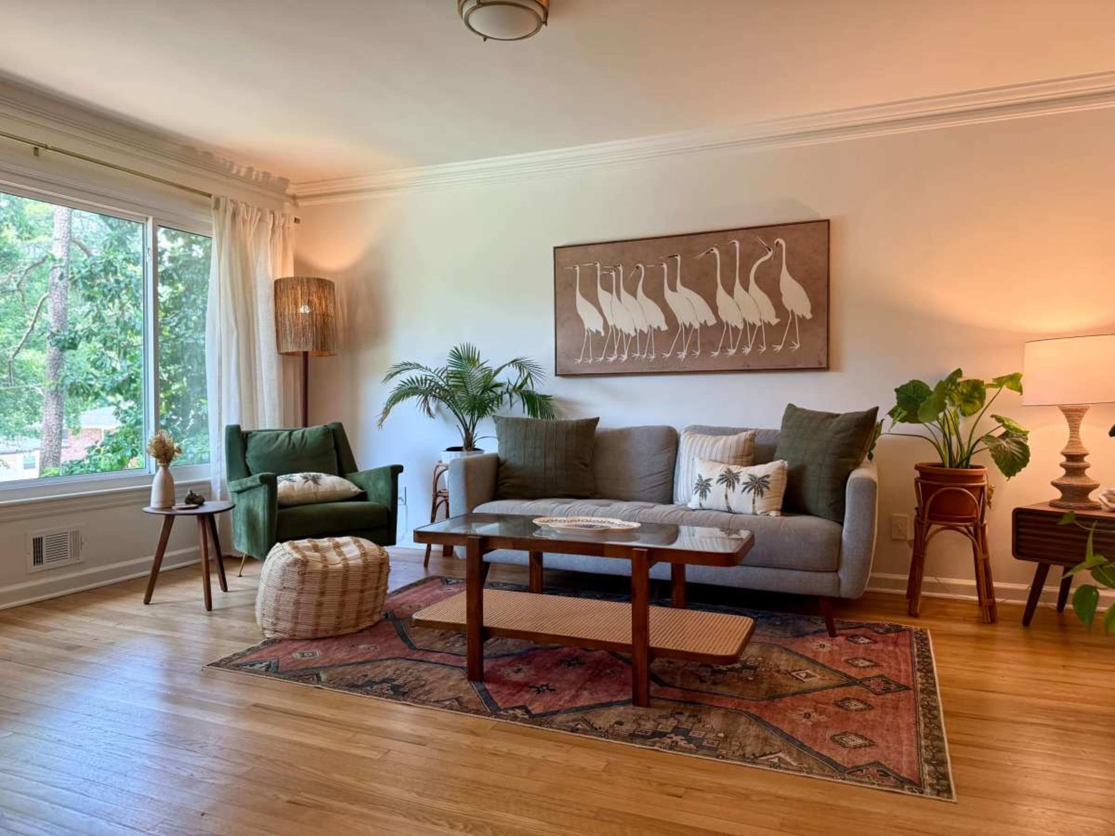 The image depicts a cozy living room with a gray sofa, a green armchair, a wooden coffee table, and decorative plants, illuminated by natural light from a large window.