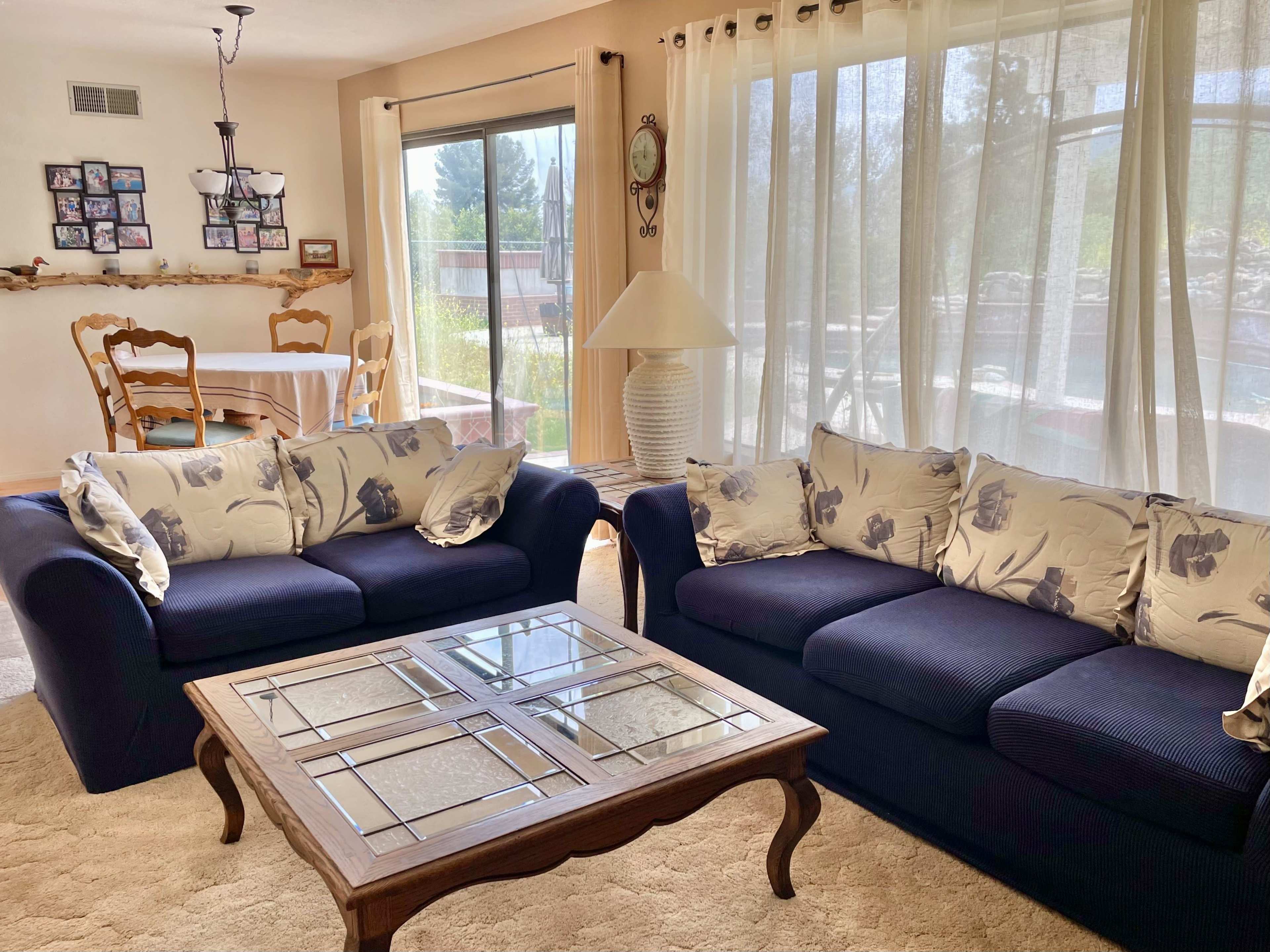 A cozy living room featuring two dark blue sofas with floral-patterned cushions, a wooden coffee table with a glass top, and large windows allowing natural light to enter.