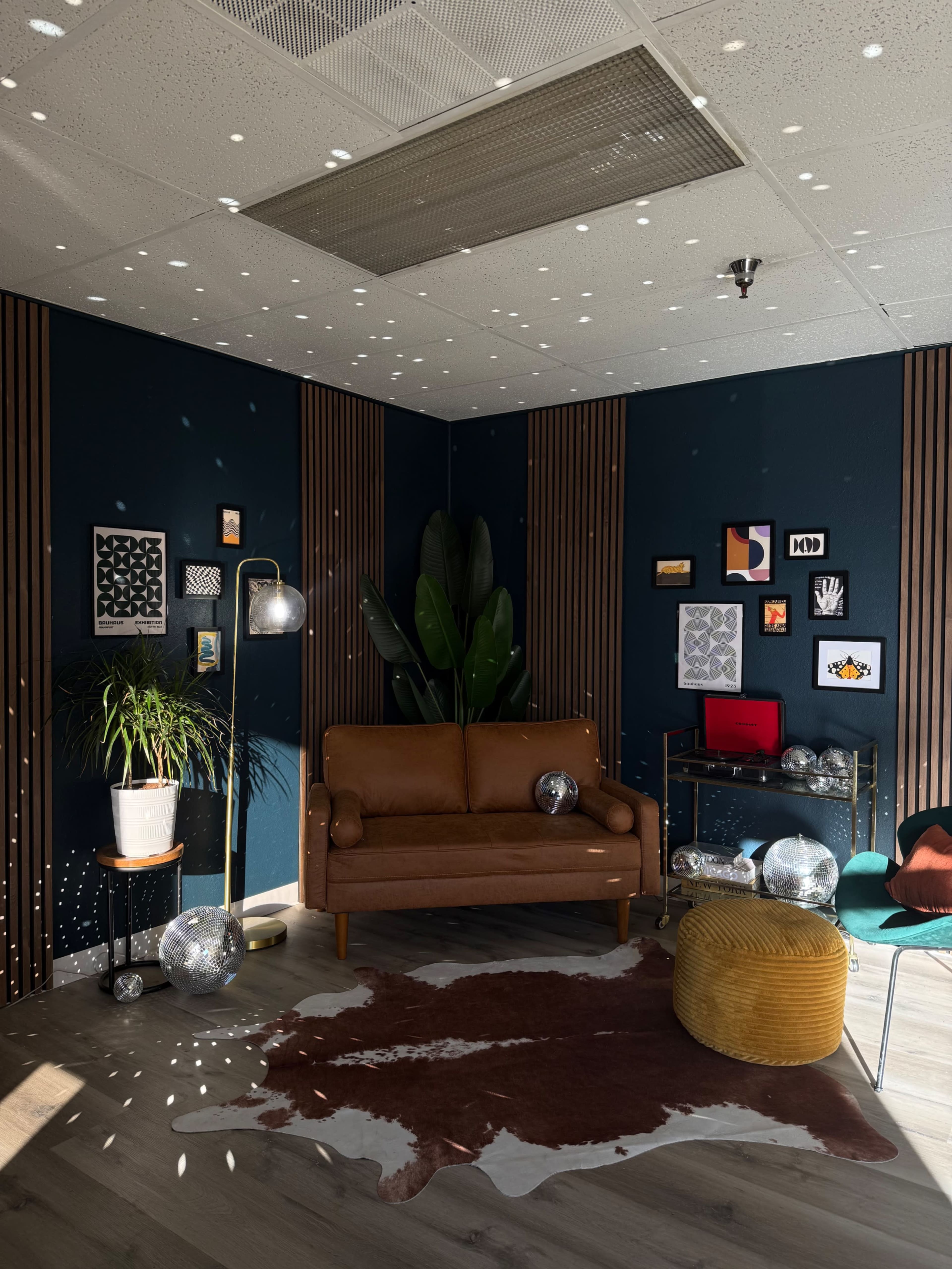 The image shows a stylish room with a brown leather couch, a green chair, various framed artworks, a potted plant, and scattered light reflections from disco balls on the walls.