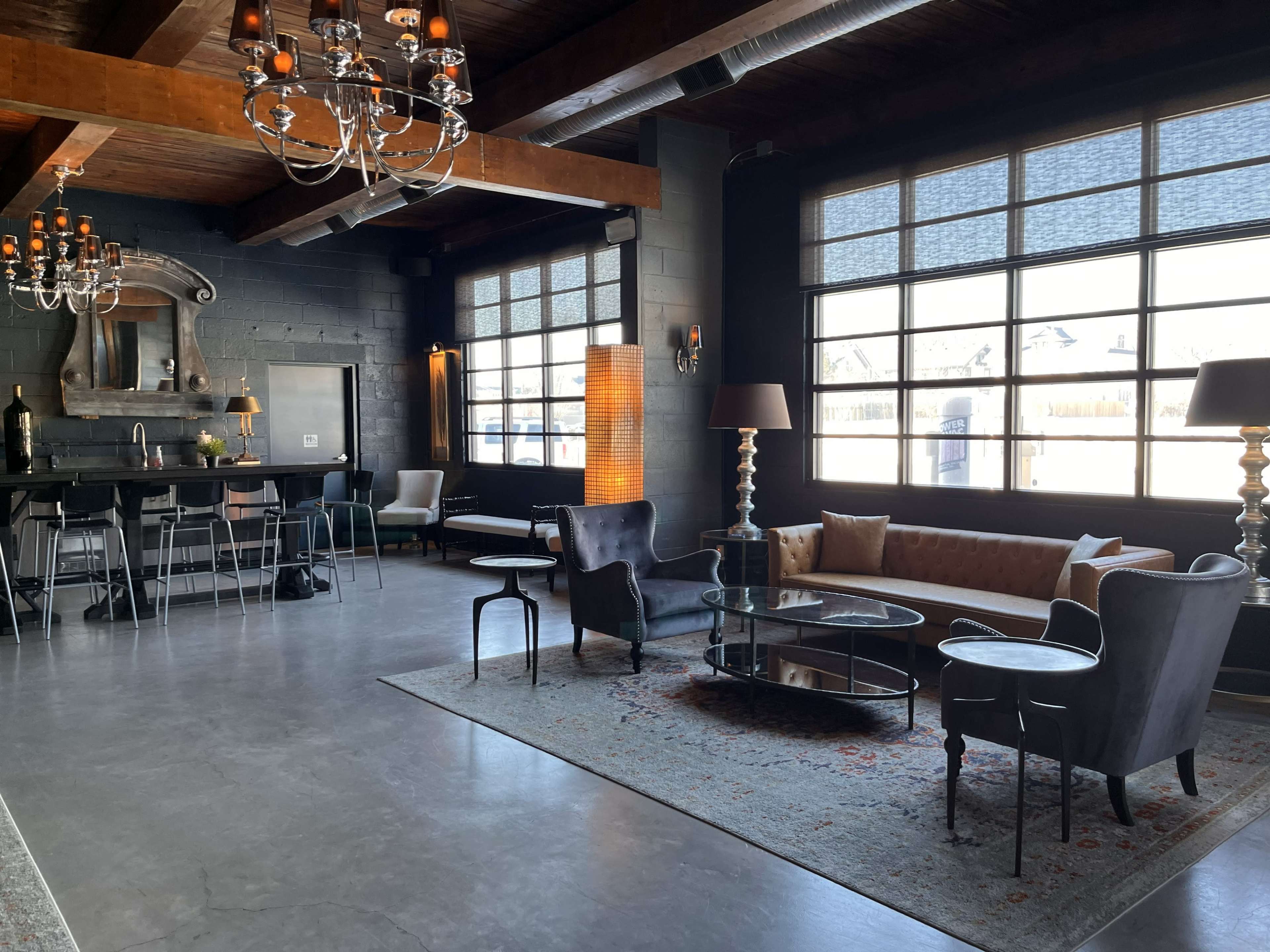 The image shows a modern lounge area with a mix of seating options, a bar in the background, and large windows allowing natural light to enter.