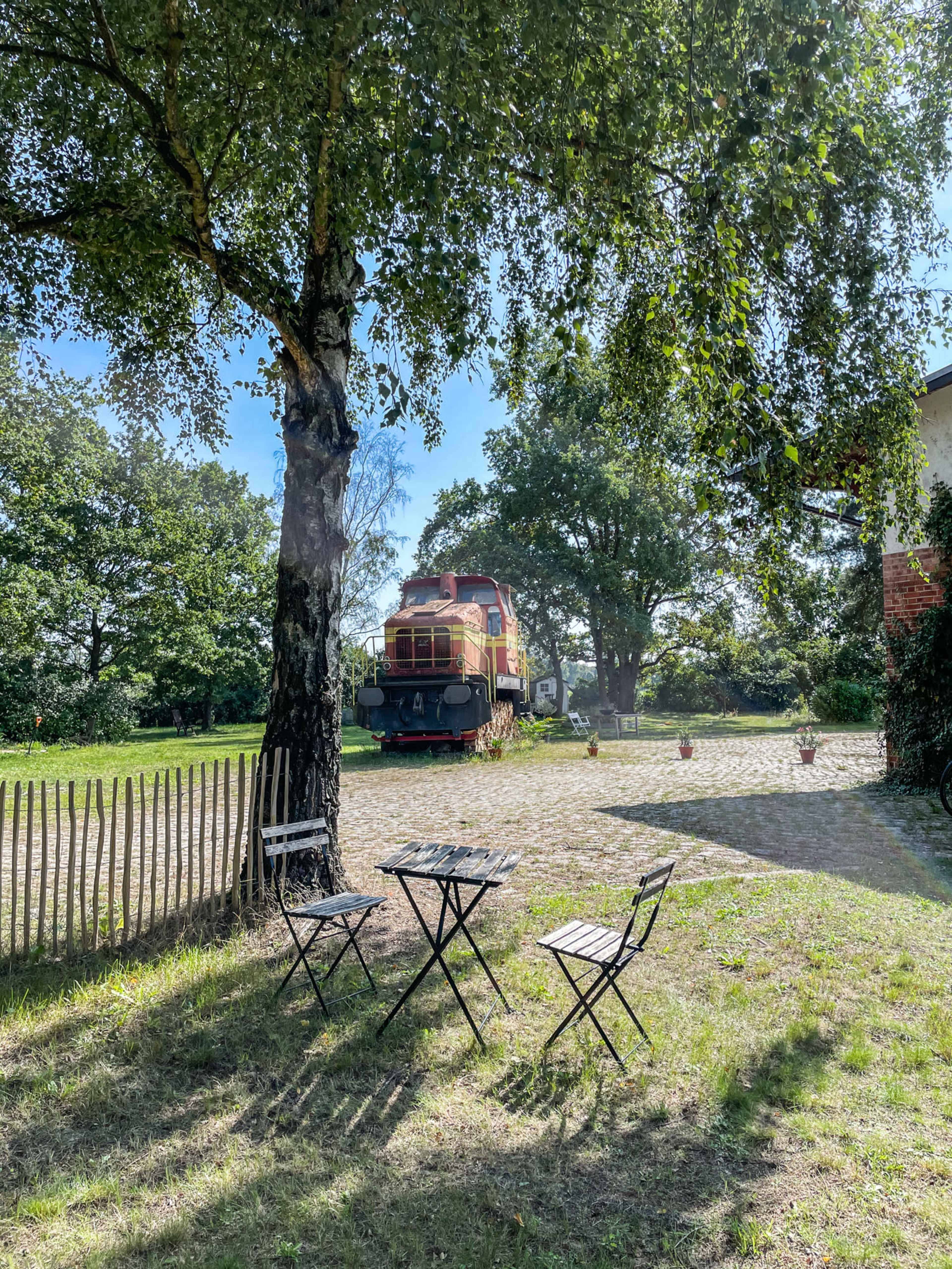A vintage train engine sits parked beside a grassy area with a small table and chairs under a tree.