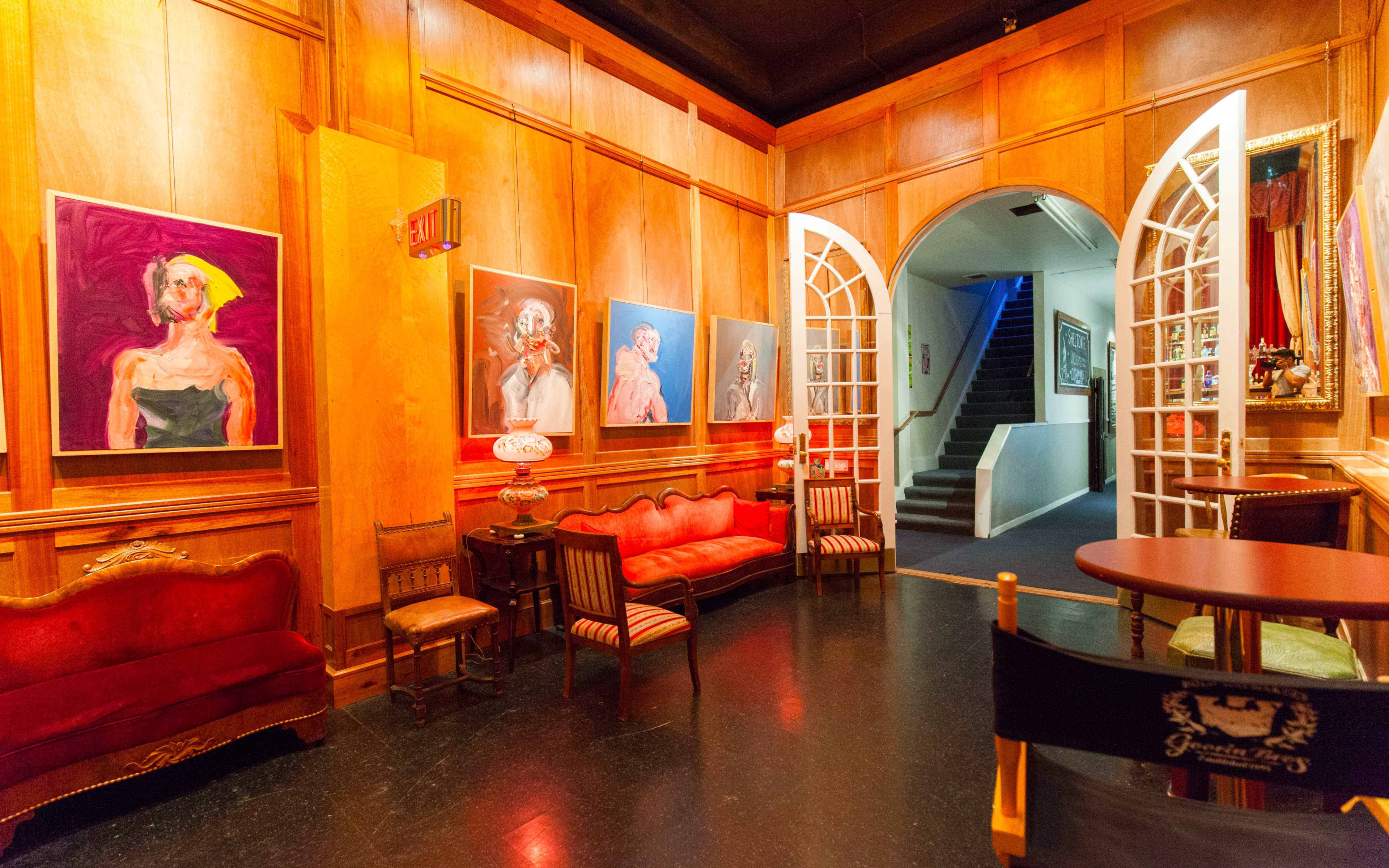 A shadowy room with wooden paneling, featuring several colorful paintings on the walls, a pair of red sofas, and a table with a director's chair near an archway leading to a staircase.