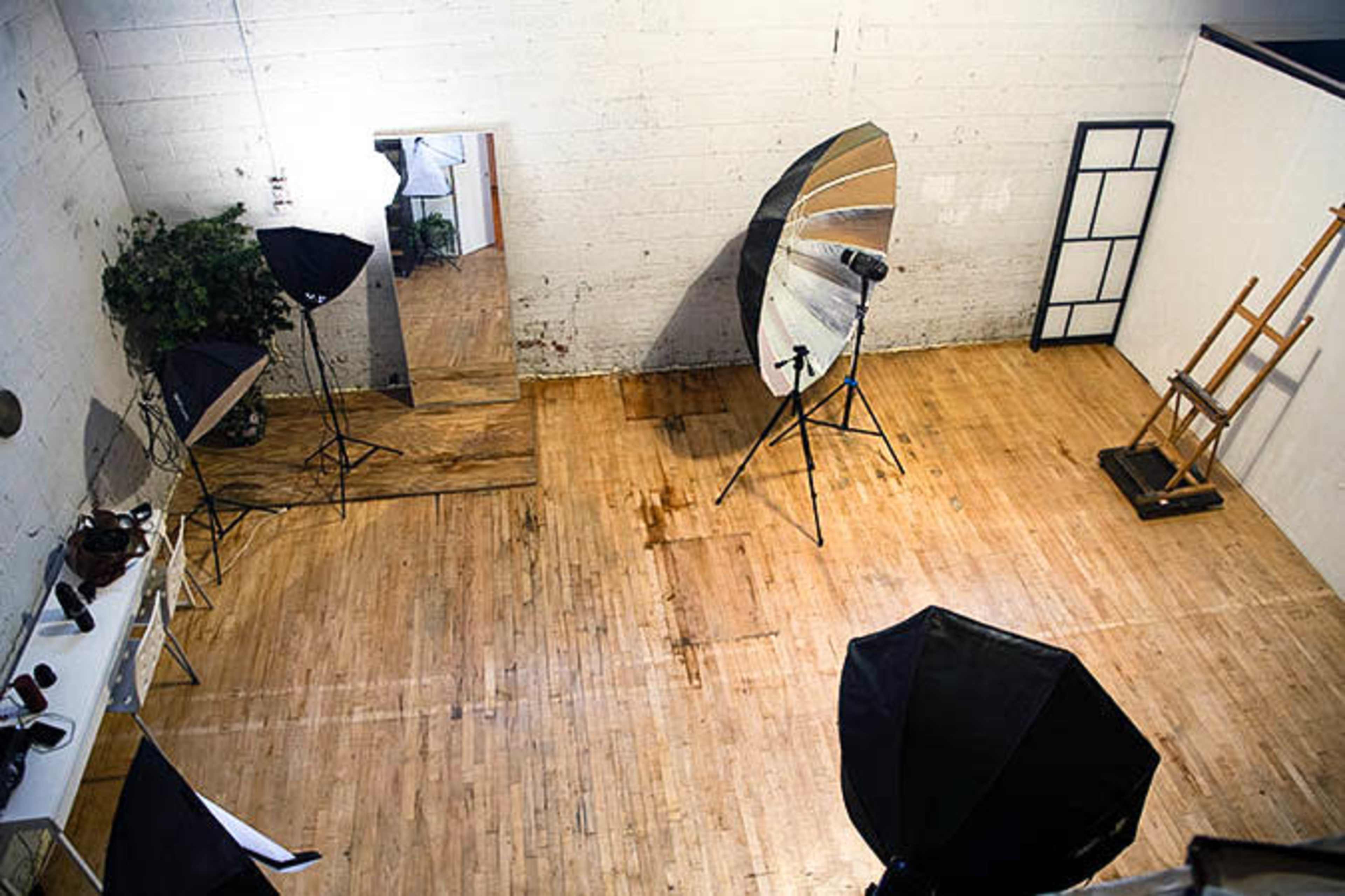 The image shows a photography studio with wooden flooring, a large mirror, various lighting equipment, and an easel against the wall.