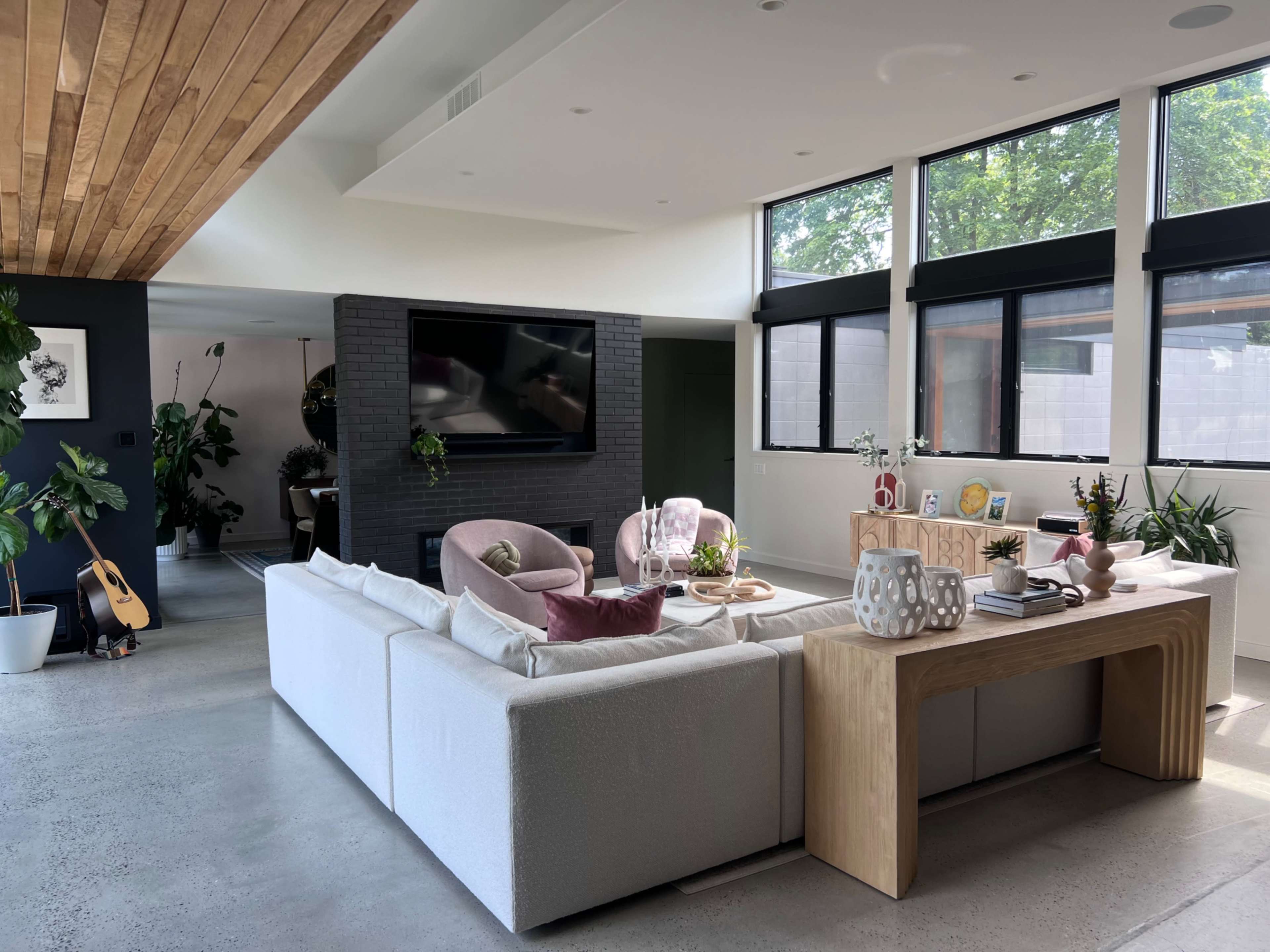 The image shows a modern living room with a sectional sofa, a wooden coffee table, large windows letting in natural light, and various plants throughout the space.