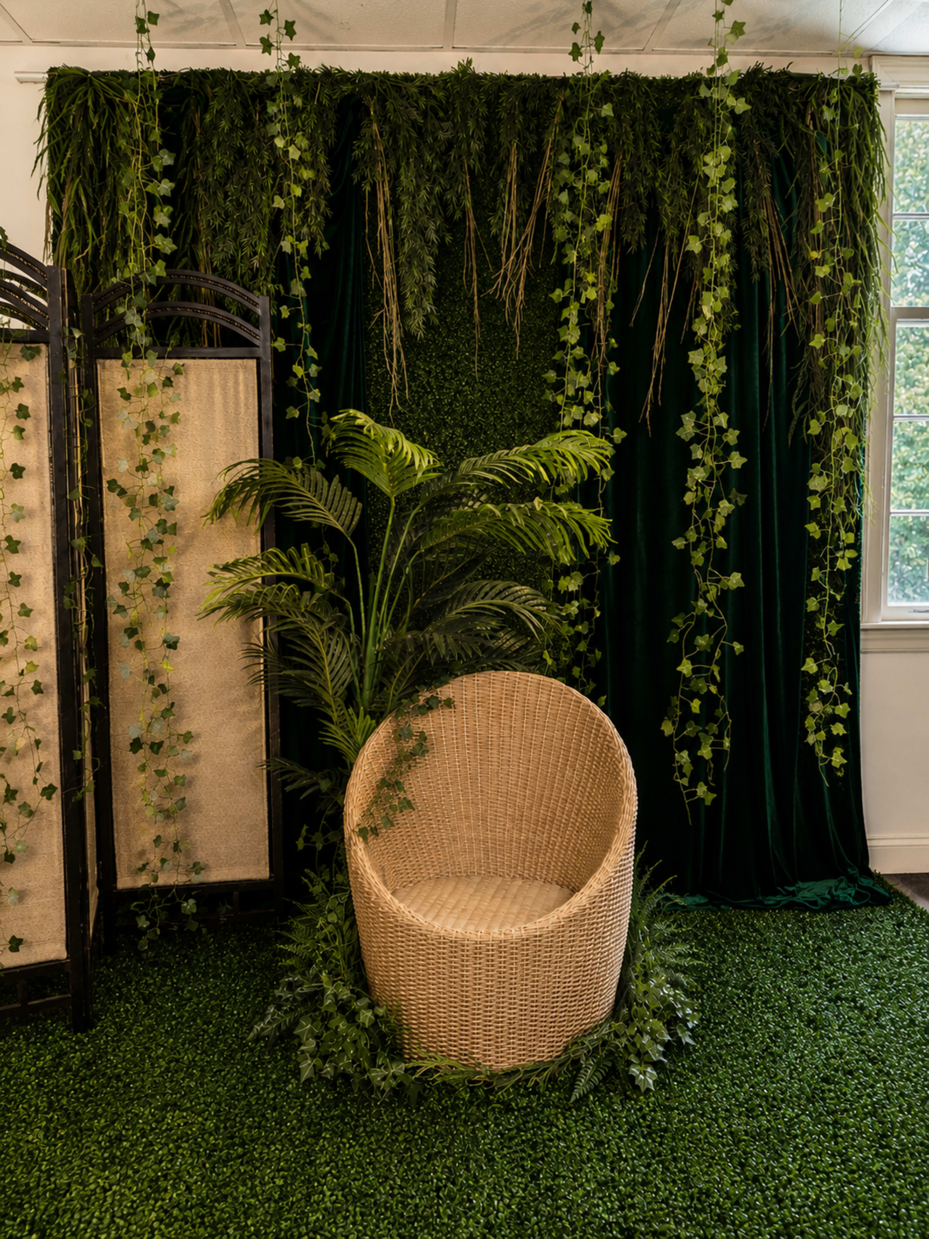 The image shows a cozy, styled corner featuring a rattan chair surrounded by lush greenery and decorative vines against a backdrop of hanging fabric and greenery.
