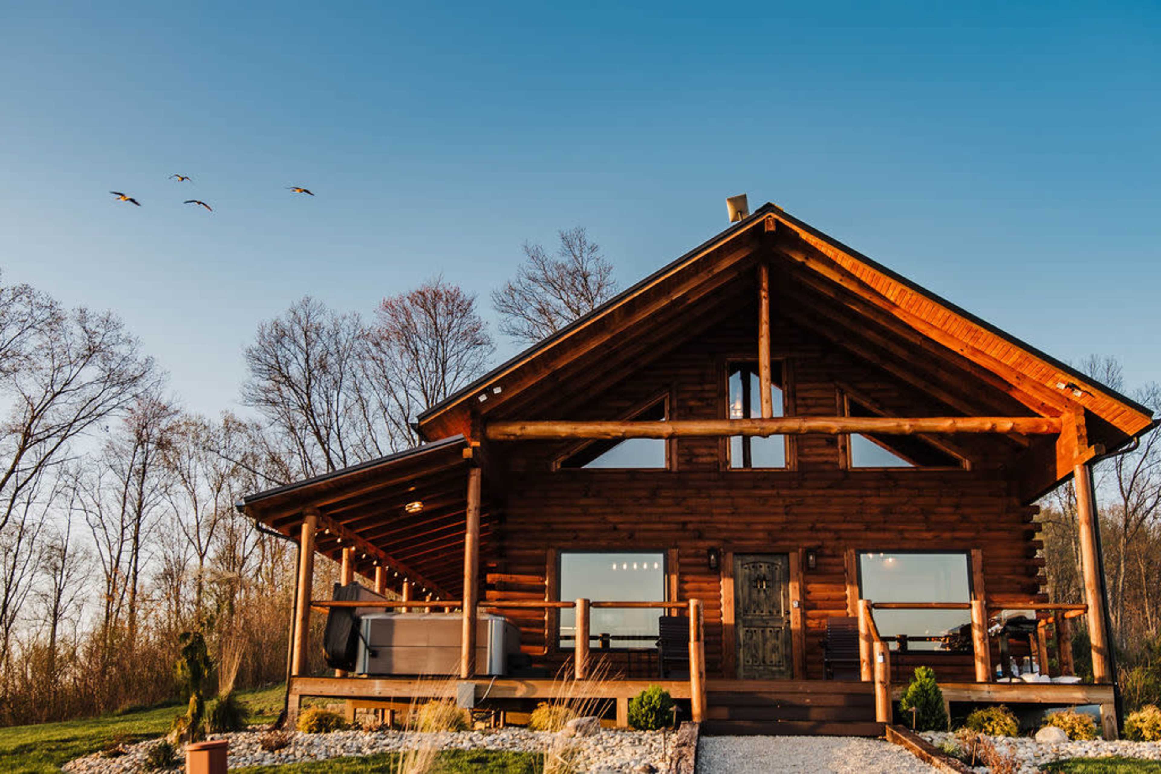 A wooden cabin with a sloped roof and large front porch is set against a clear blue sky, with several birds flying overhead and trees in the background.