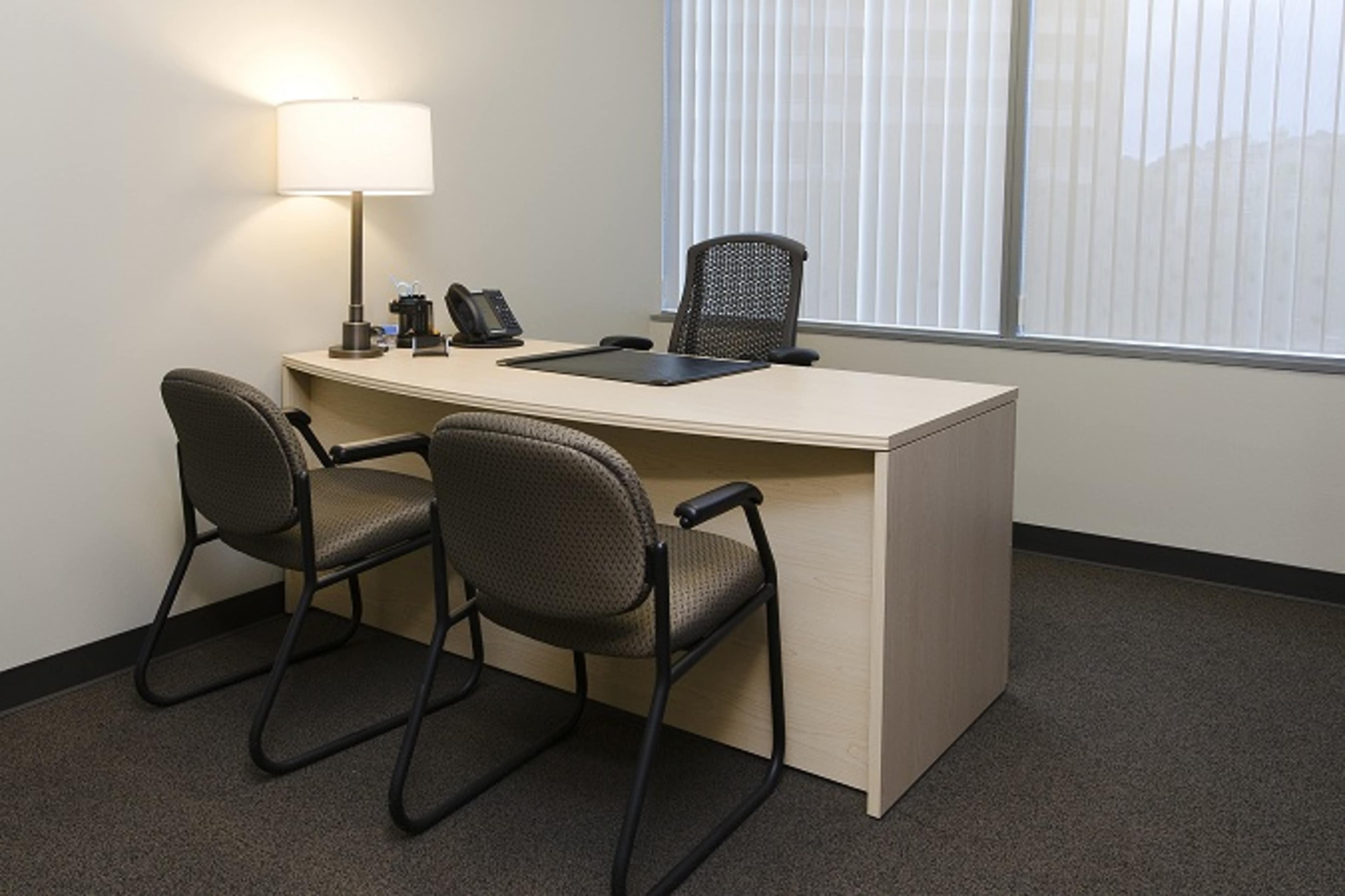The image shows a minimalist office space featuring a desk with a lamp, phone, and two chairs adjacent to it.