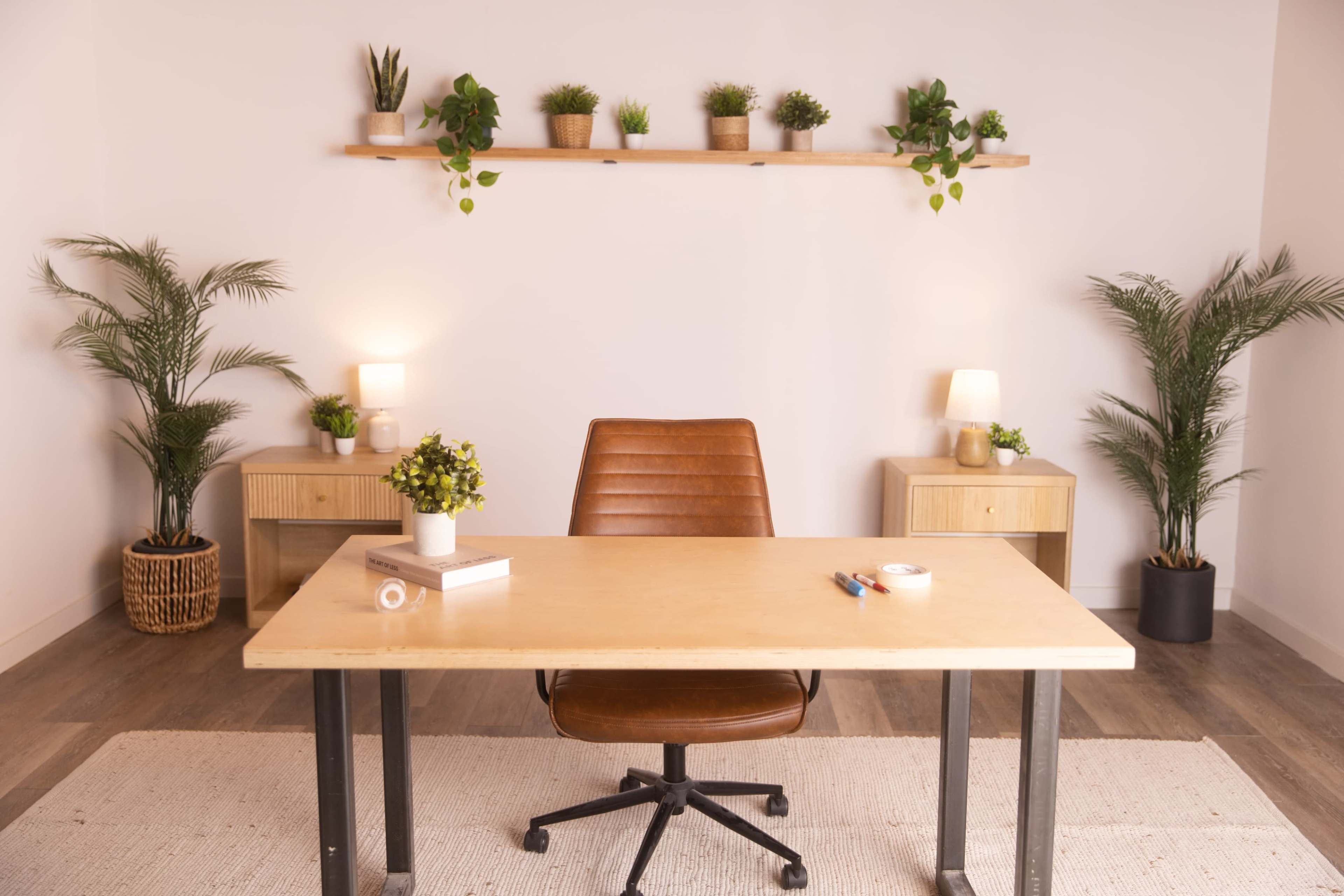 A simple office space features a wooden desk with a brown chair, flanked by two cabinets and adorned with potted plants on a shelf and the floor.