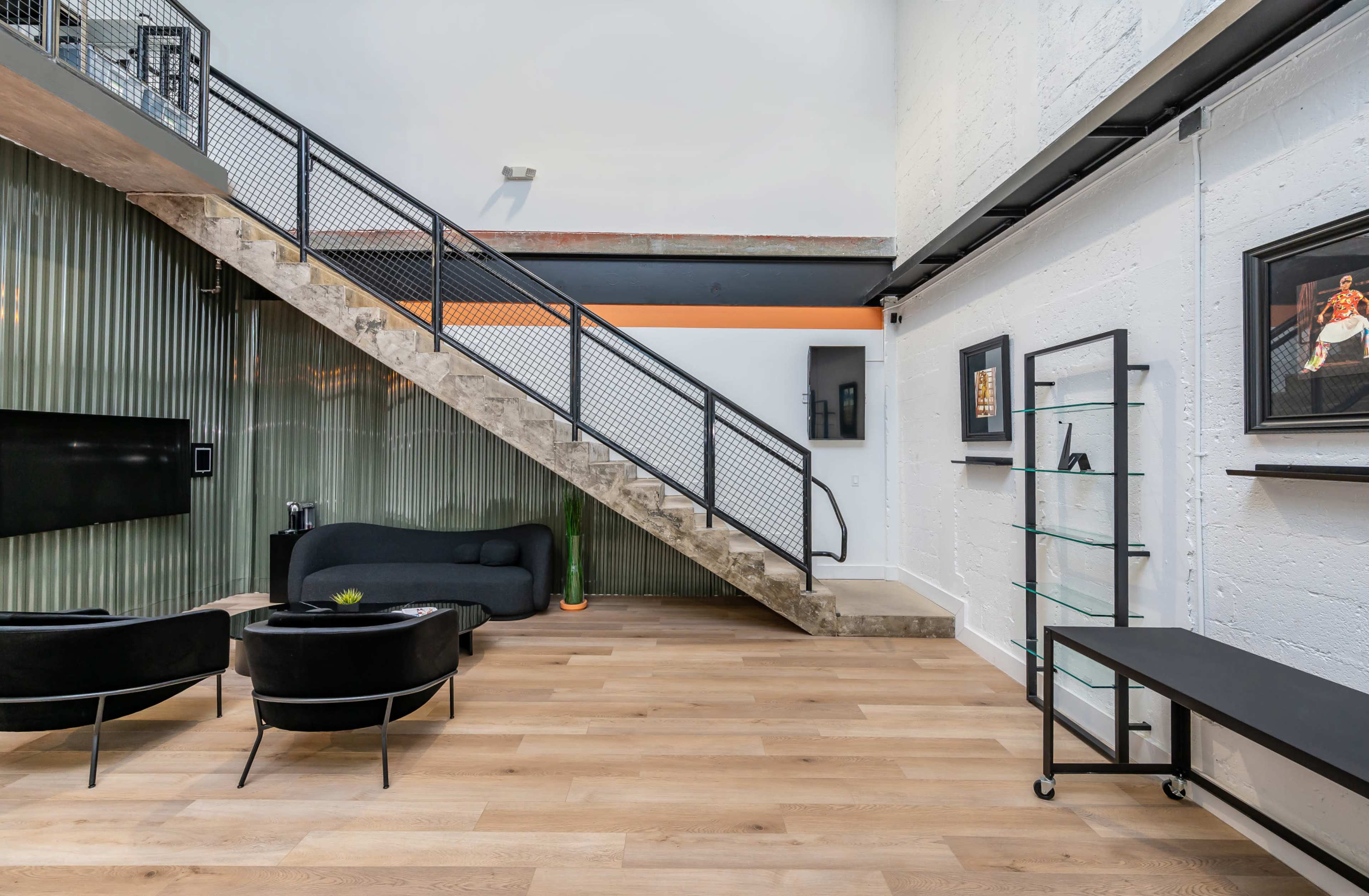 A spacious interior features a modern staircase, a black sofa, and minimalist furniture elements against a backdrop of wooden flooring and textured walls.
