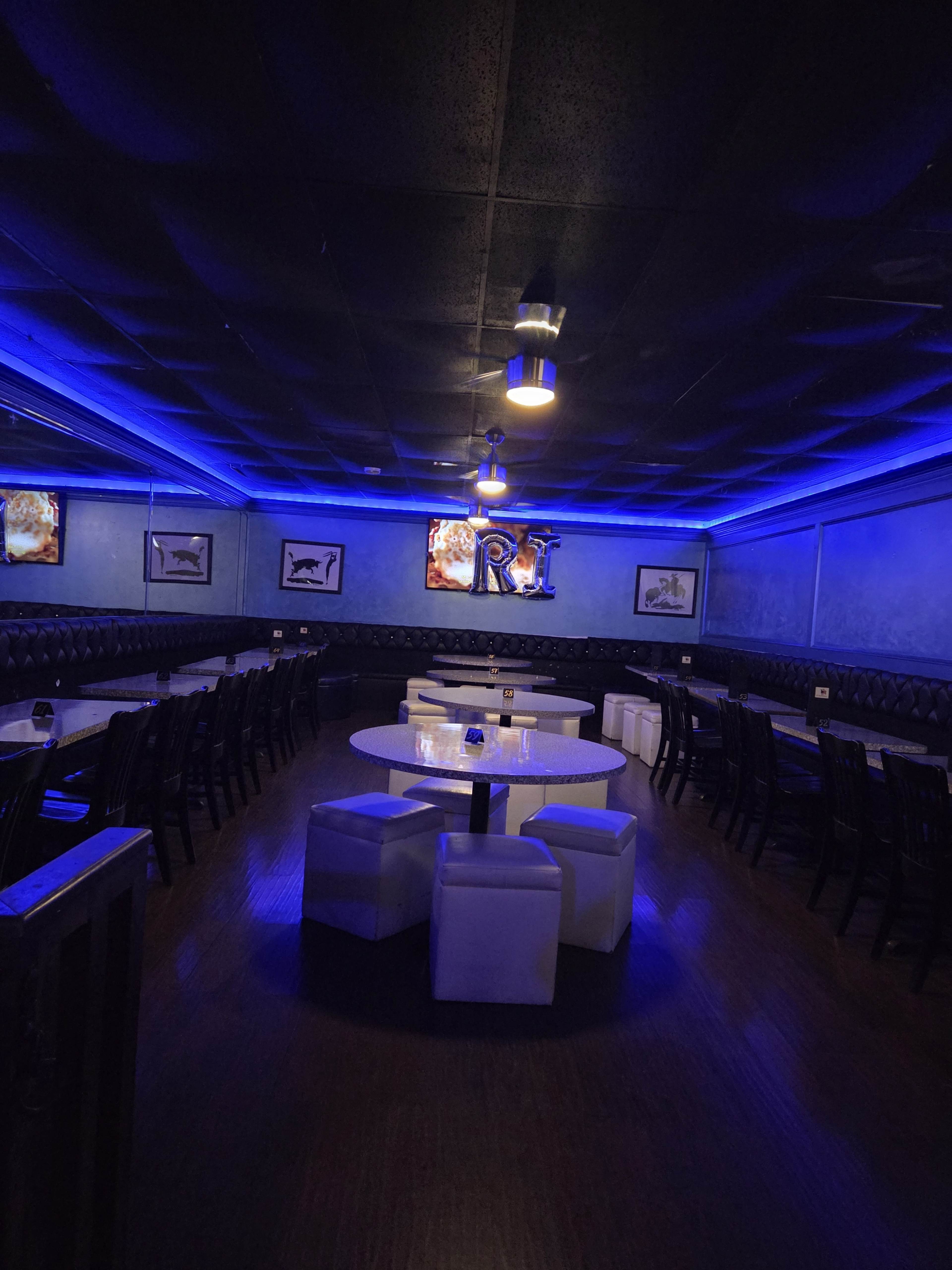 The image shows a dimly lit restaurant interior featuring blue lighting, several round tables with white seating, and framed pictures on the walls.