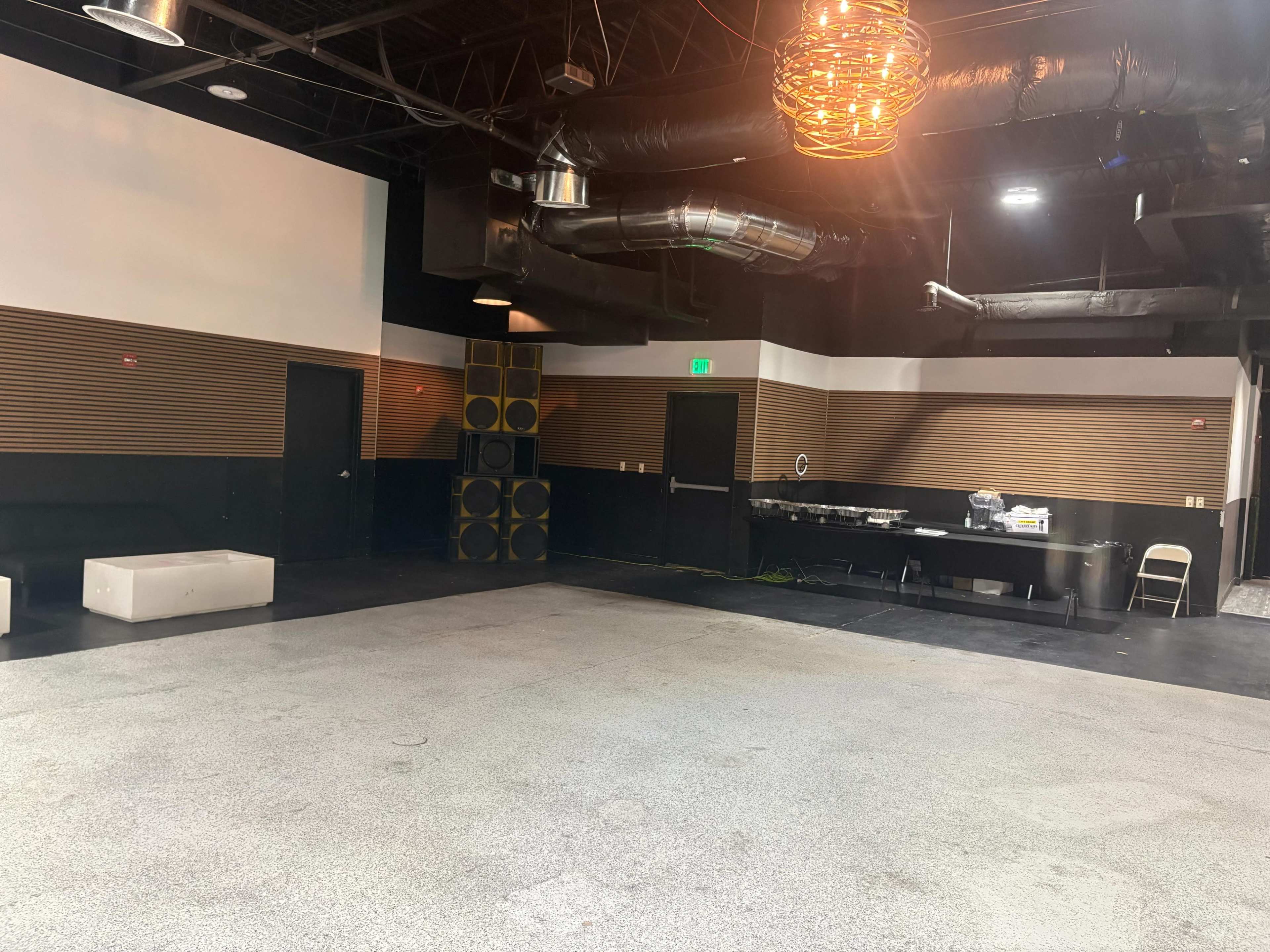 The image shows an empty indoor space with a polished concrete floor, wooden wall panels, and minimal furnishings, including a few chairs and a sound system setup in the corner.