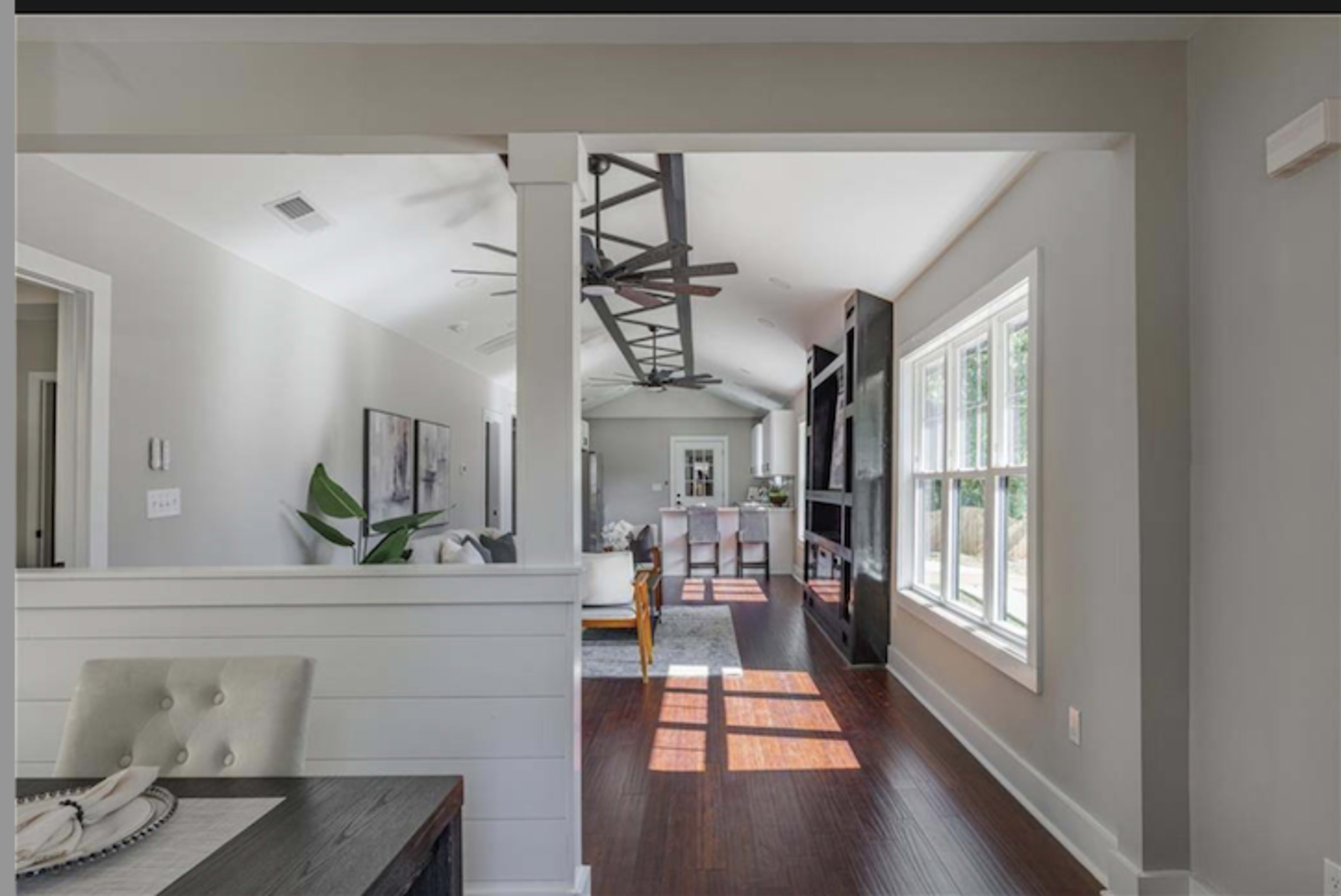 The image shows a bright, open interior space of a modern home with a living area, dining area, and large windows.