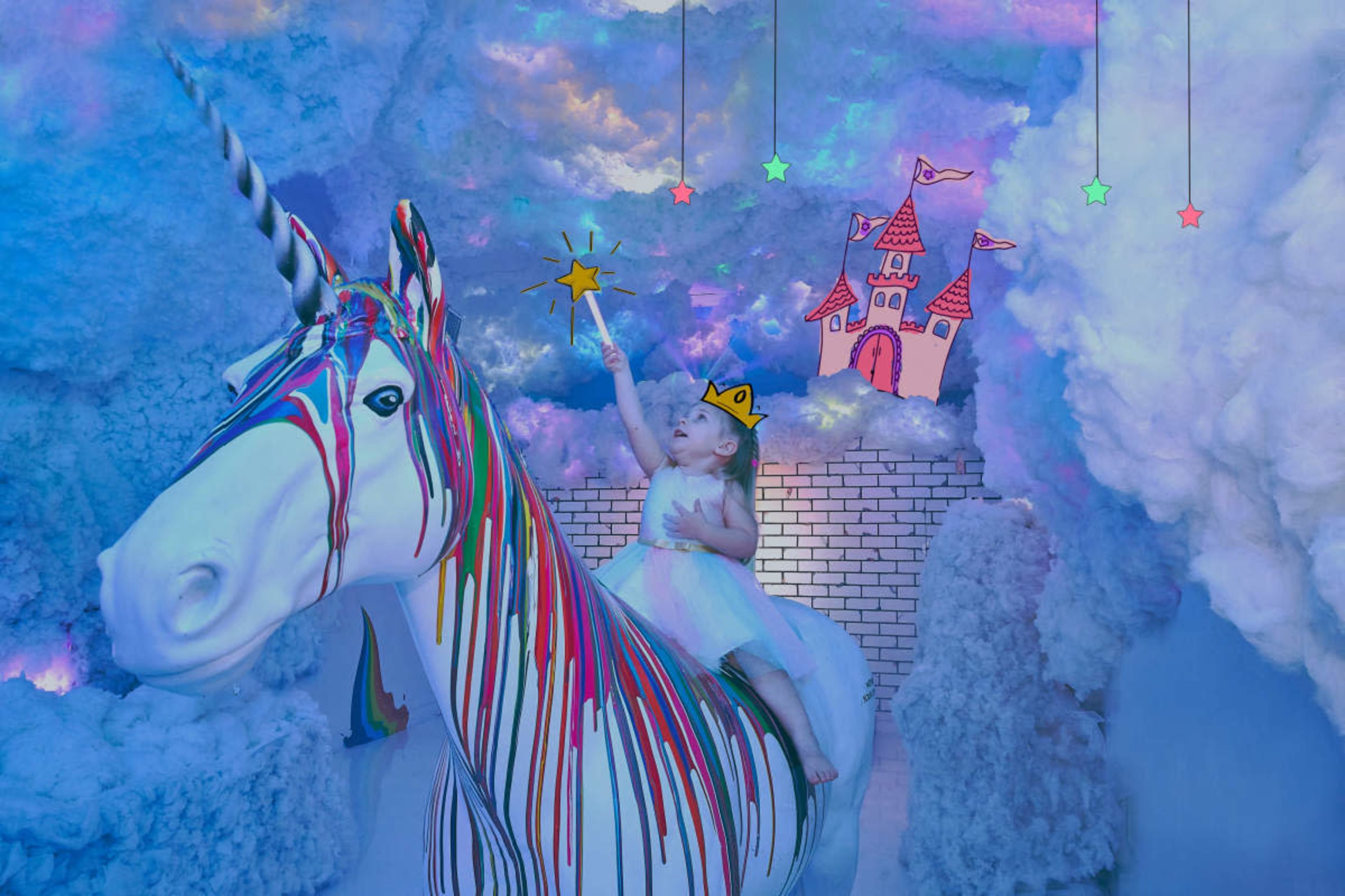 A child wearing a crown sits on a colorful unicorn statue in a whimsical, cloud-filled setting.
