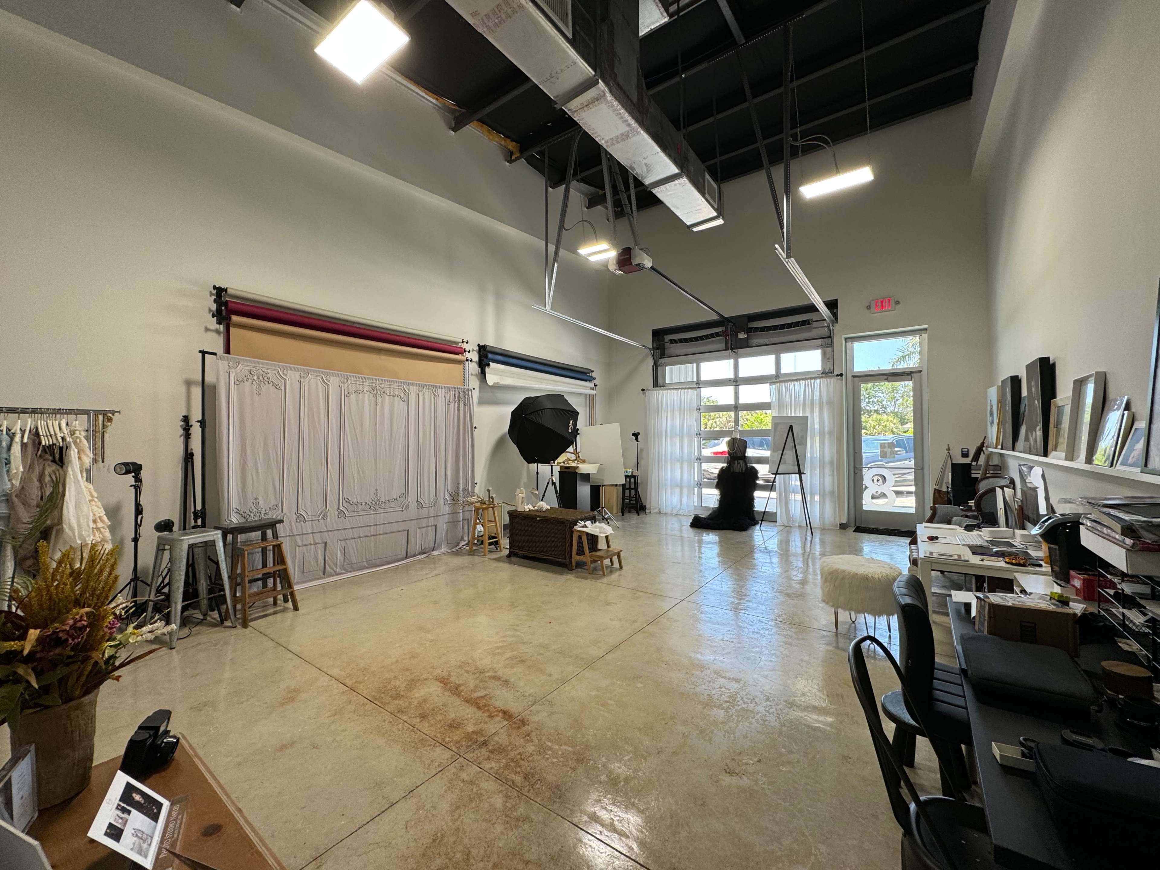 A spacious studio with a backdrop setup, a large light umbrella, various furniture pieces, and a view of the outside through glass doors.