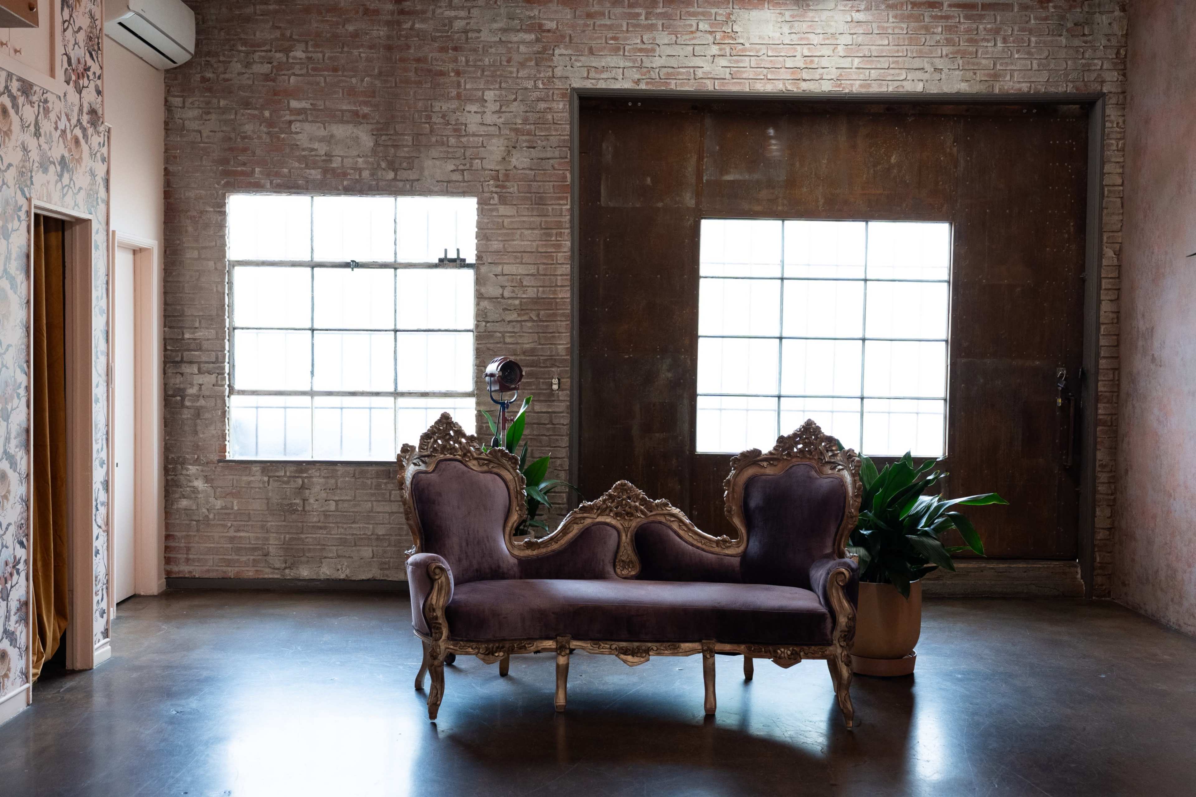 The image shows a vintage-style purple couch positioned in an industrial space with exposed brick walls and large windows.