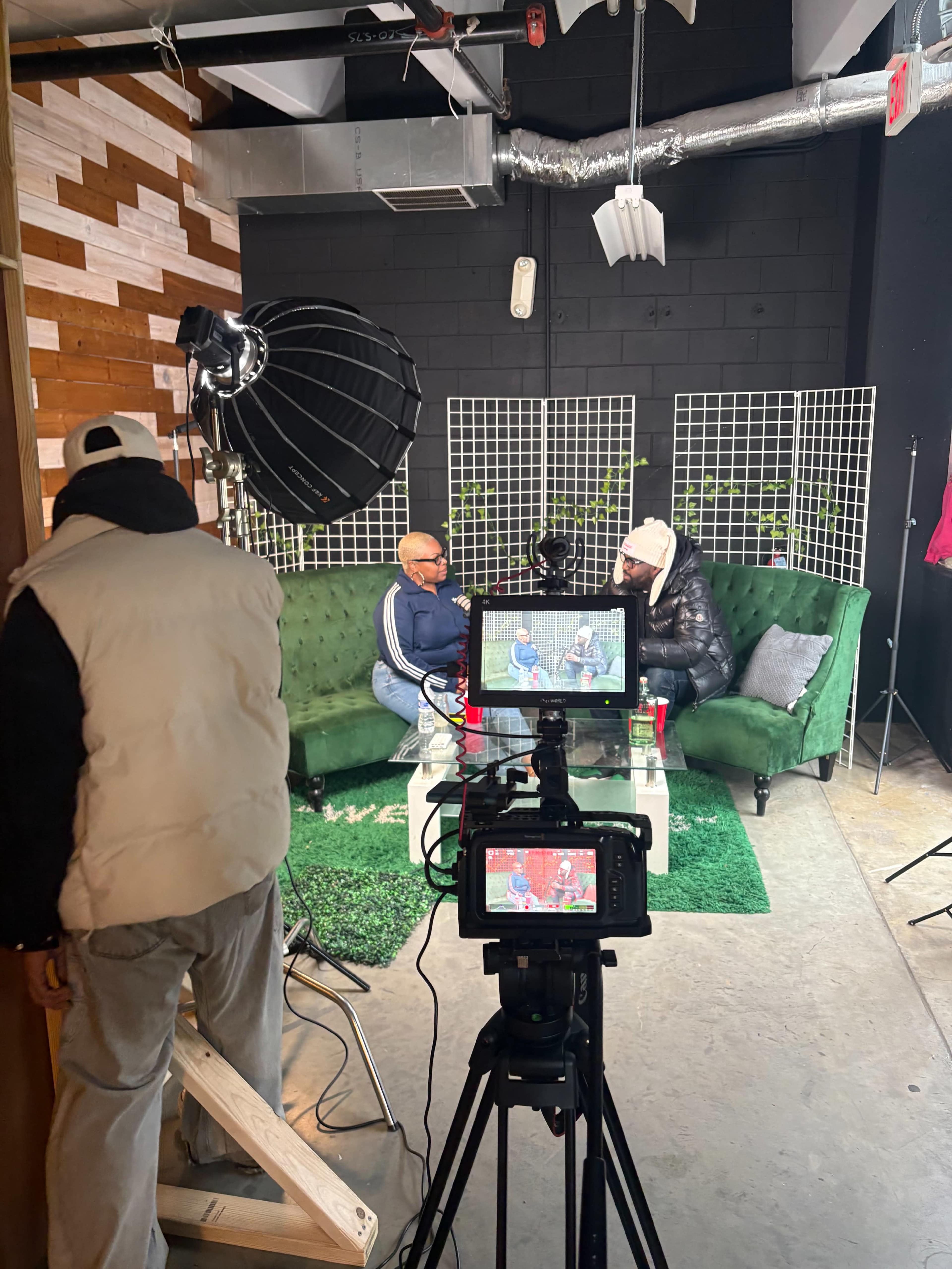 The image shows two individuals seated on a green couch in a studio setting, while a camera and lighting equipment are positioned nearby.