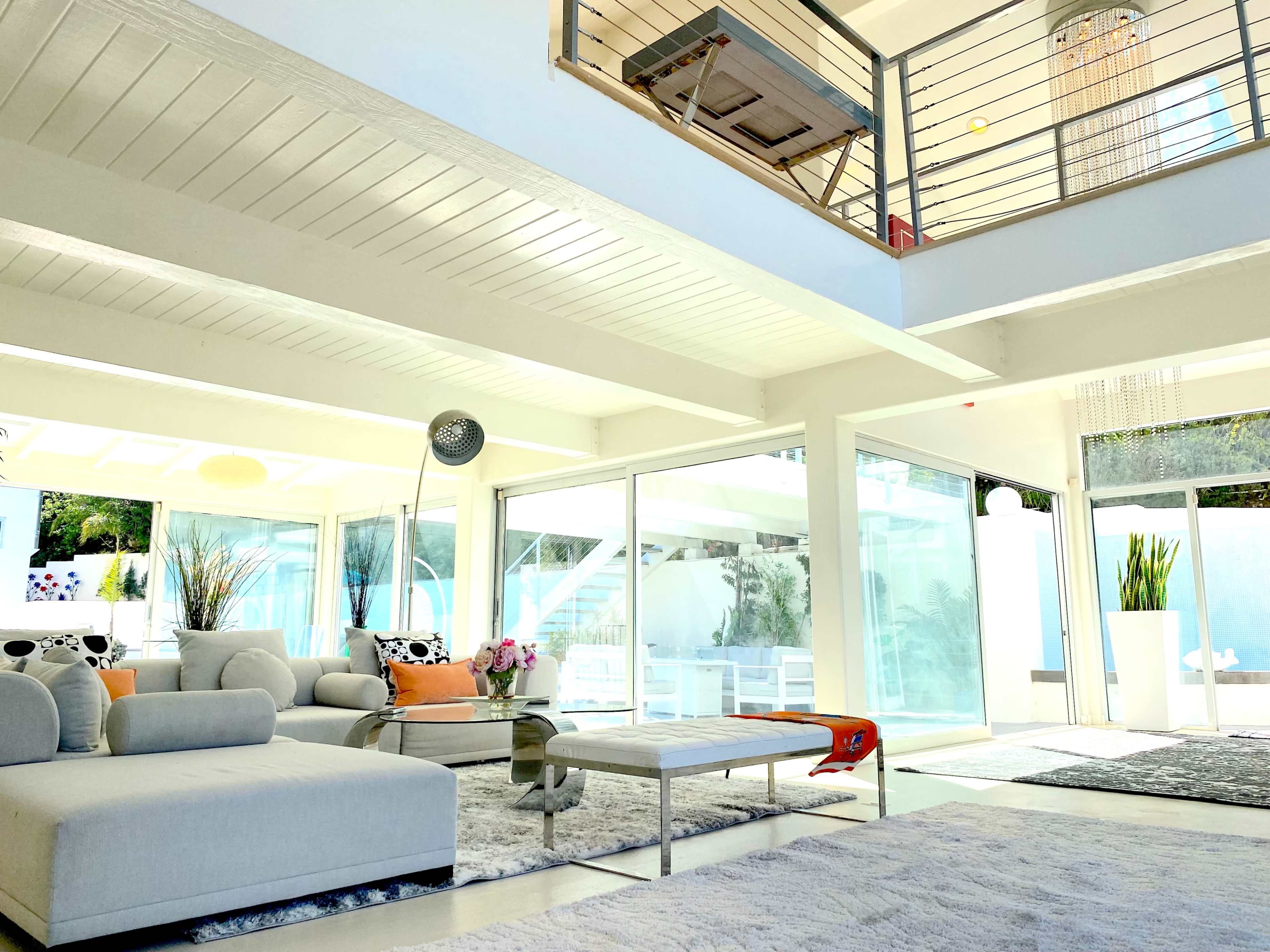 The image shows a modern living room with large glass doors, a white sectional sofa, and a coffee table, illuminated by natural light.