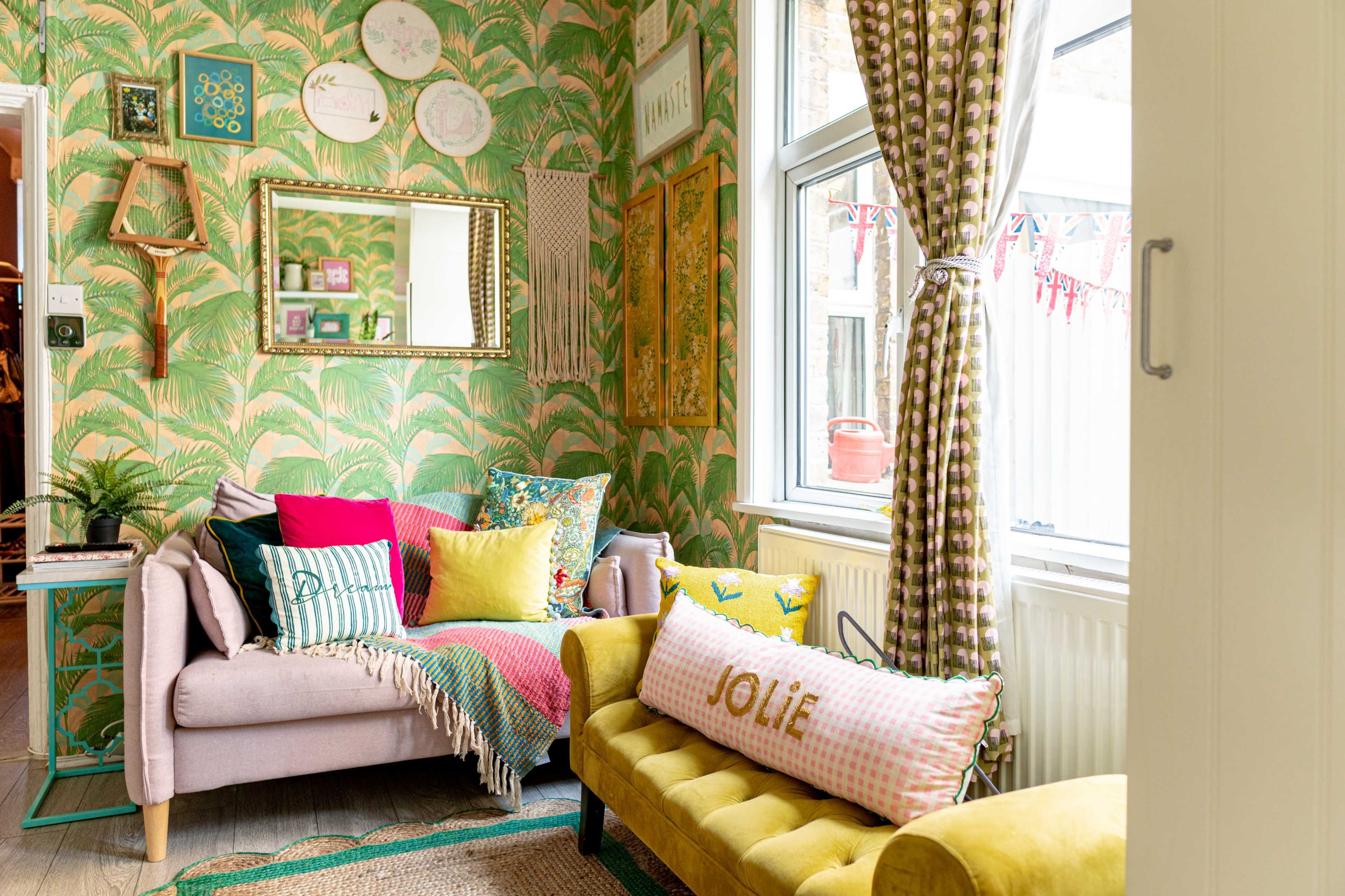 The image shows a colorful living room with patterned wallpaper, two sofas adorned with decorative pillows, and a large window allowing natural light to enter.