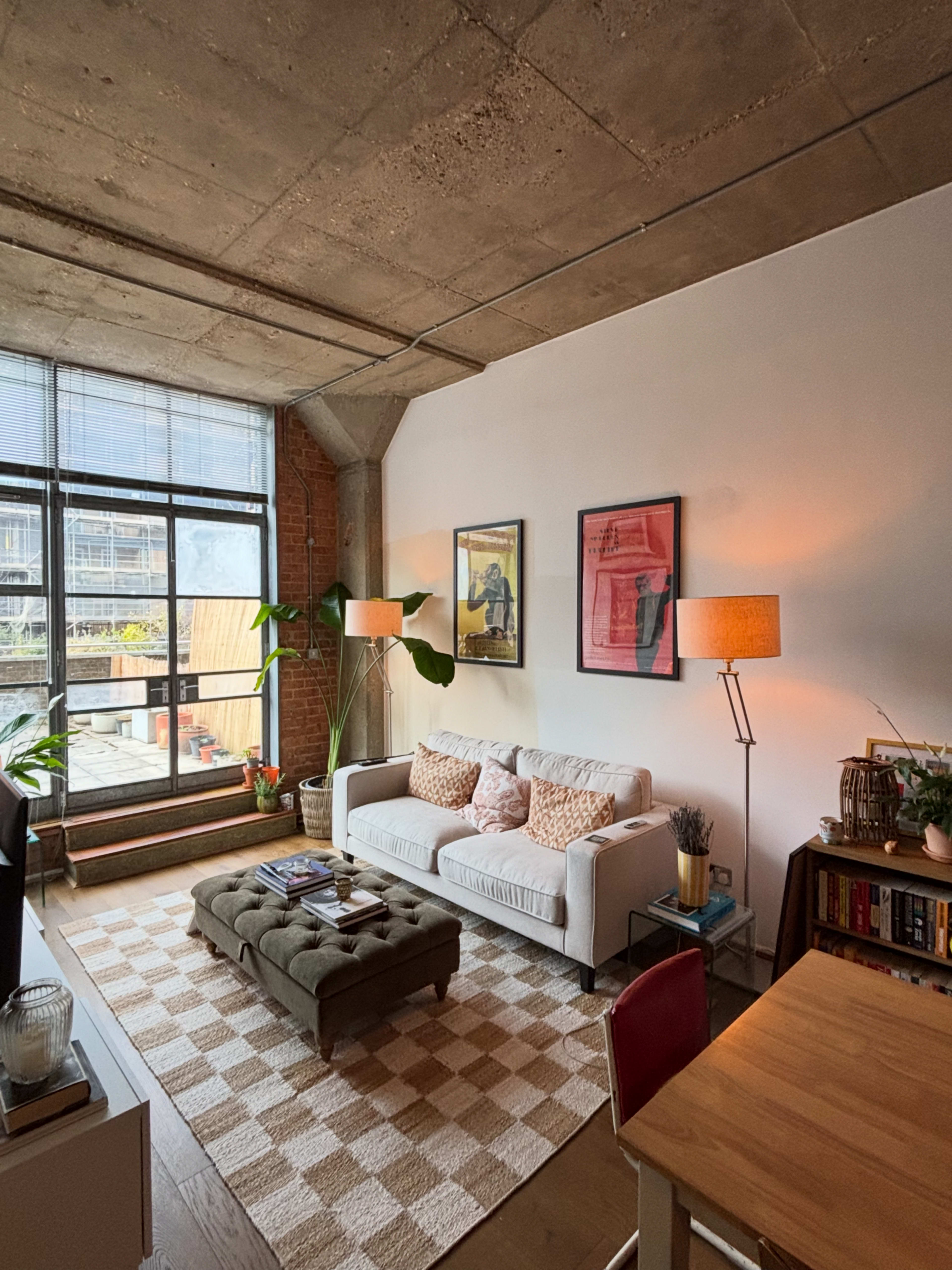 The image depicts a modern living room with a white sofa, a coffee table, a dining area, and large windows allowing natural light to enter.