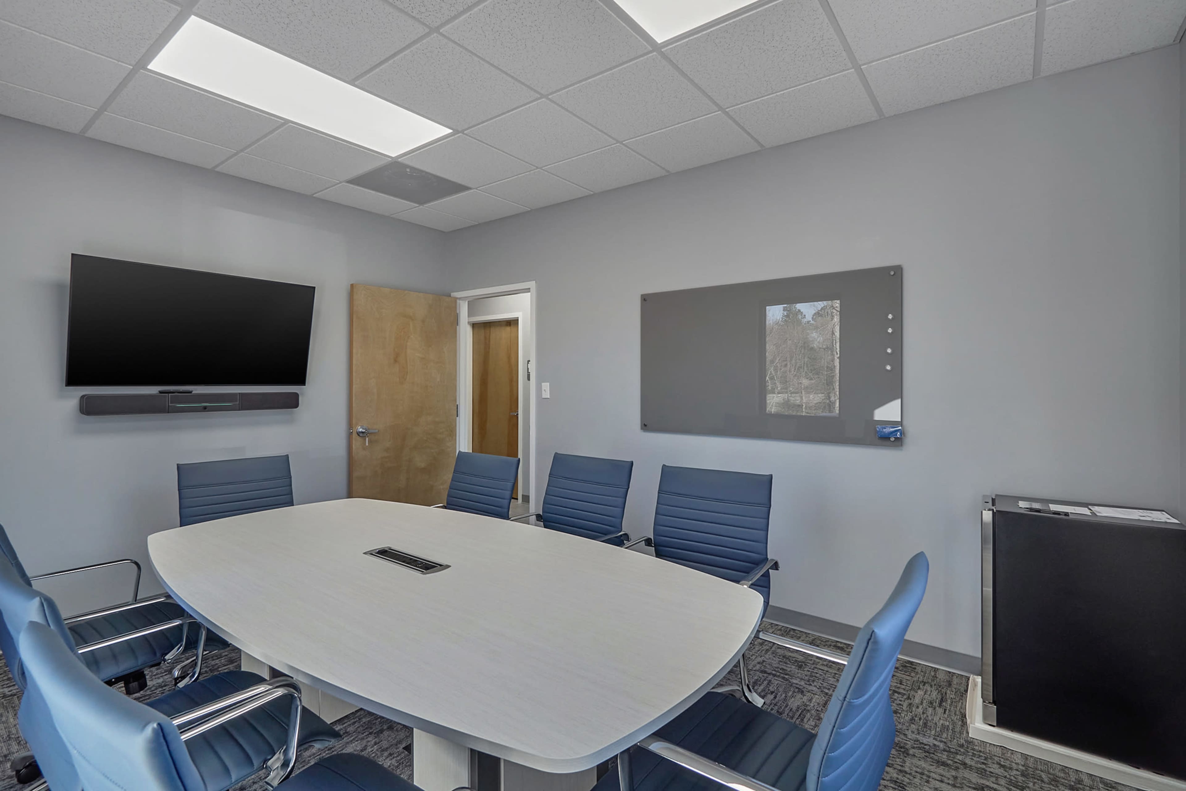 A conference room with a large oval table surrounded by several blue chairs, a television mounted on the wall, and a dry erase board.