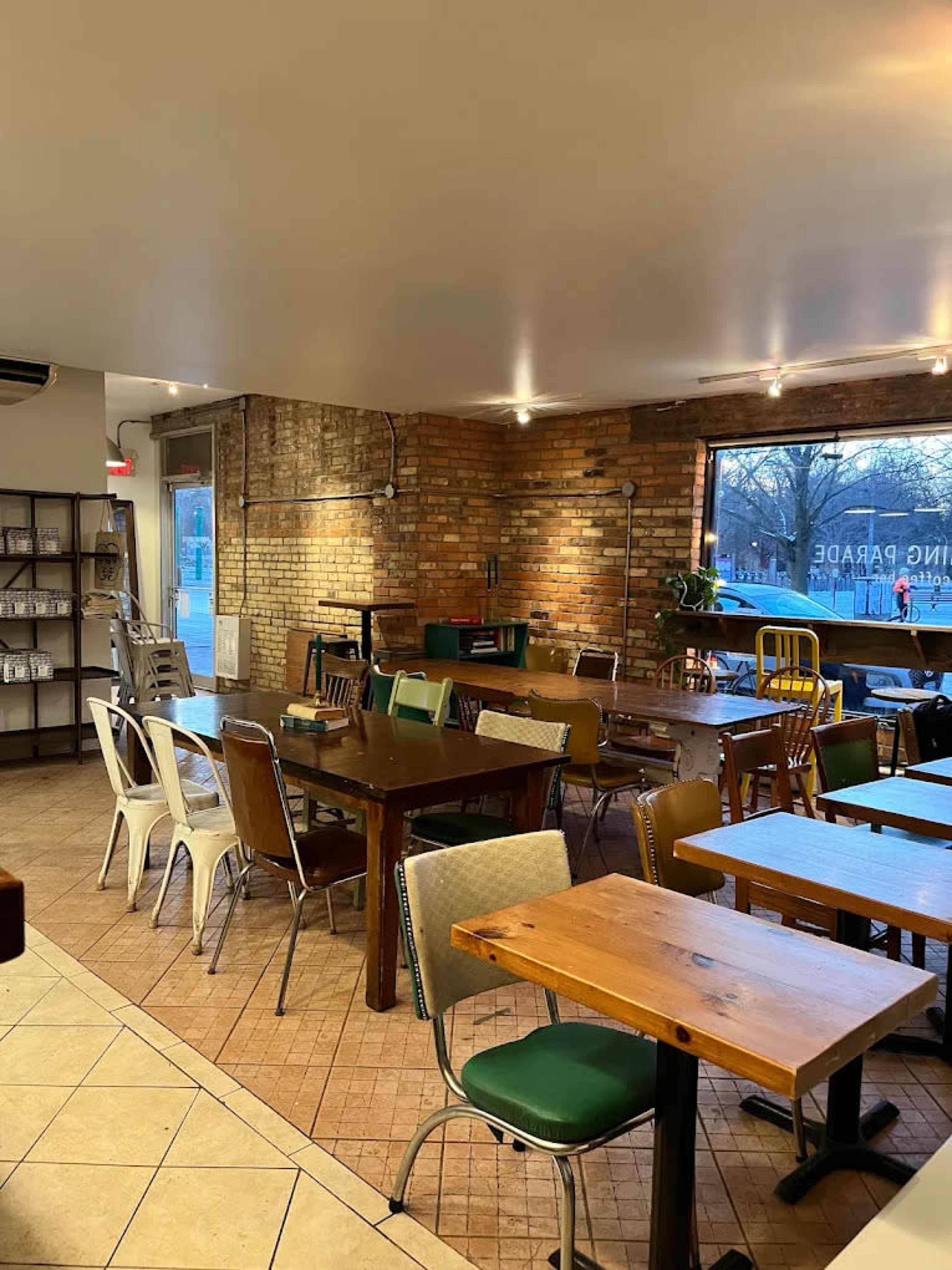 The image shows an empty restaurant interior featuring various tables and chairs, exposed brick walls, and large windows.