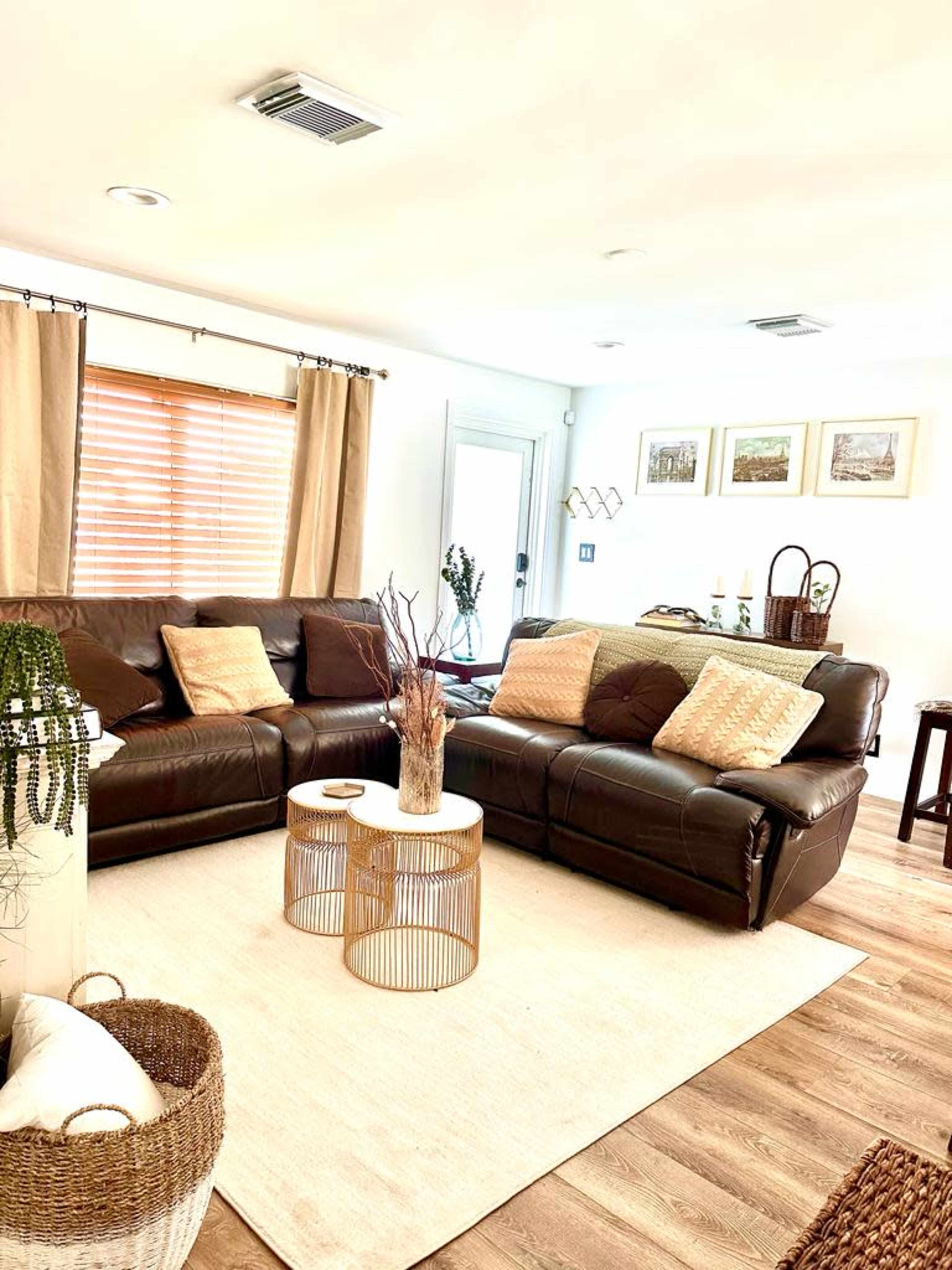The living room features a brown leather sectional sofa, a round coffee table, and neutral-colored decor with window treatments and wall art.