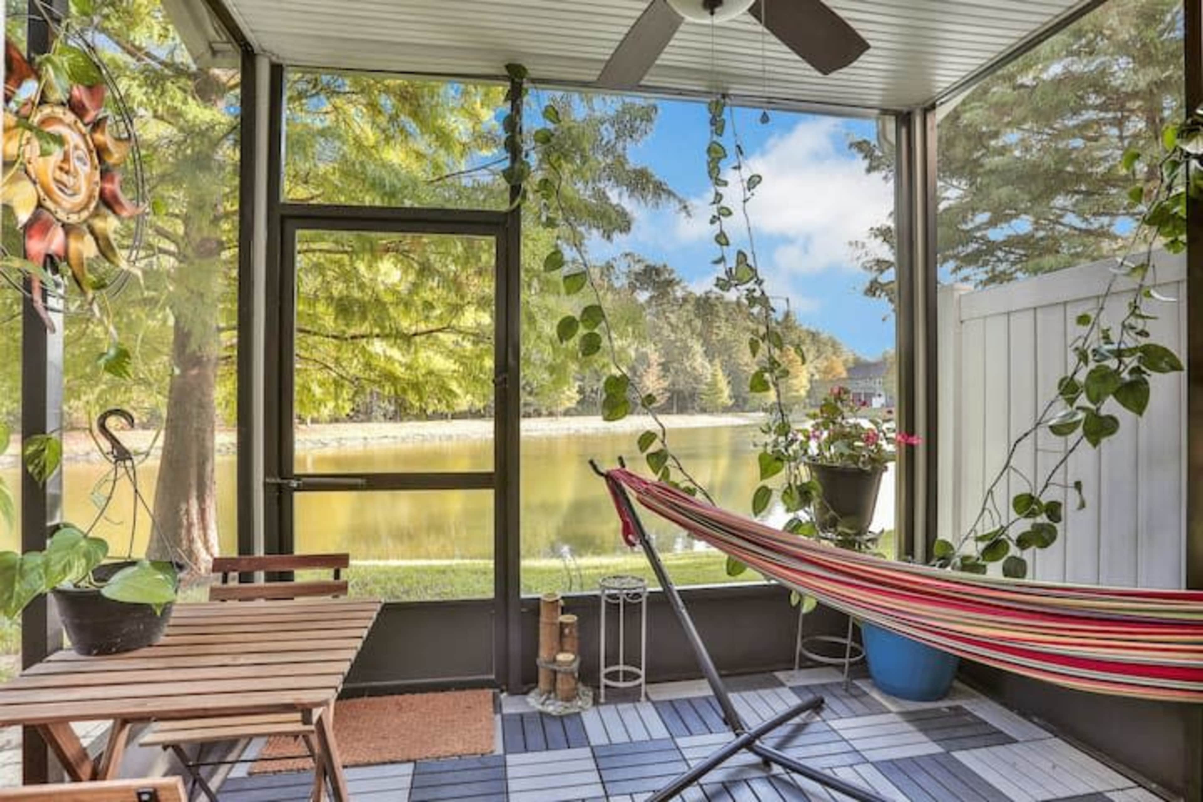 A screened-in porch features a hammock, a small wooden table, and plants, overlooking a serene lake.