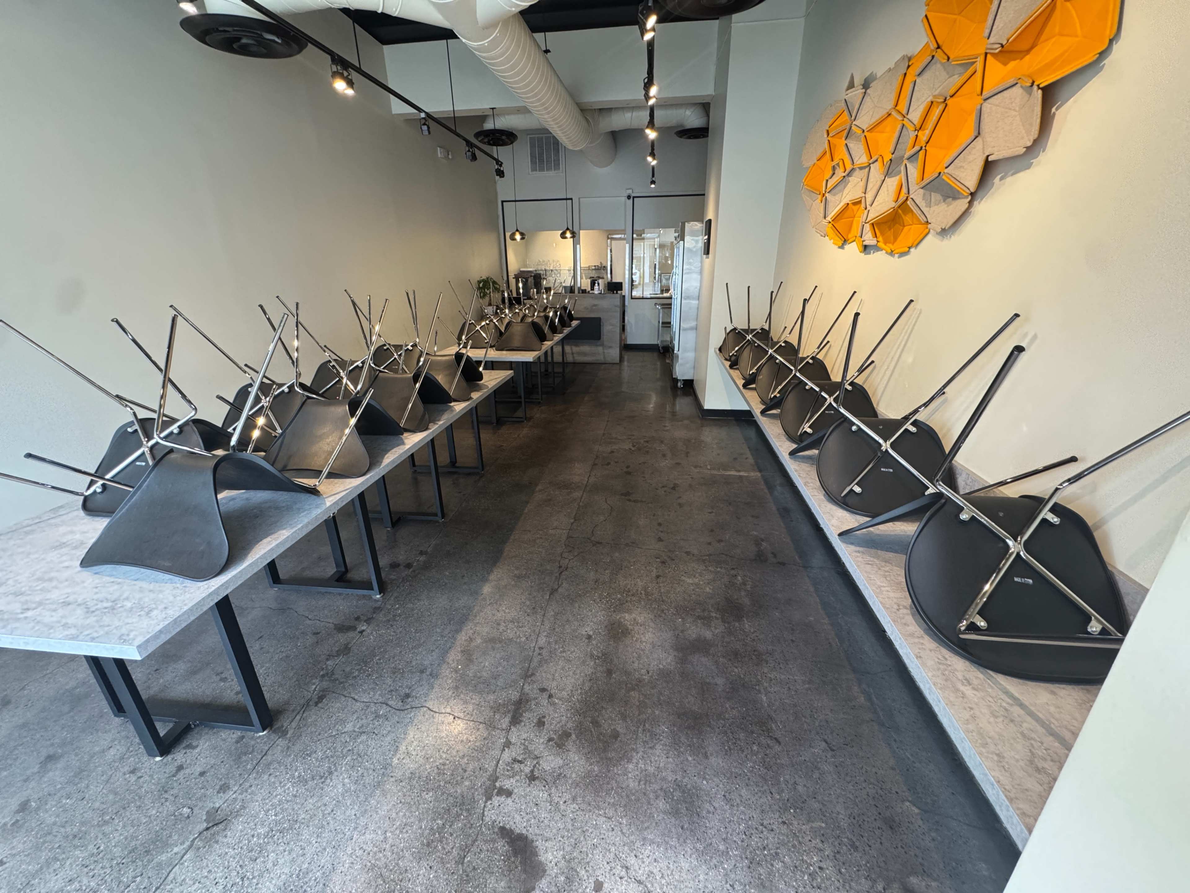 The image shows a clean, minimalist cafe or restaurant interior with metal and wooden tables, where all the chairs are stacked upside down on the tables, leaving the space empty.