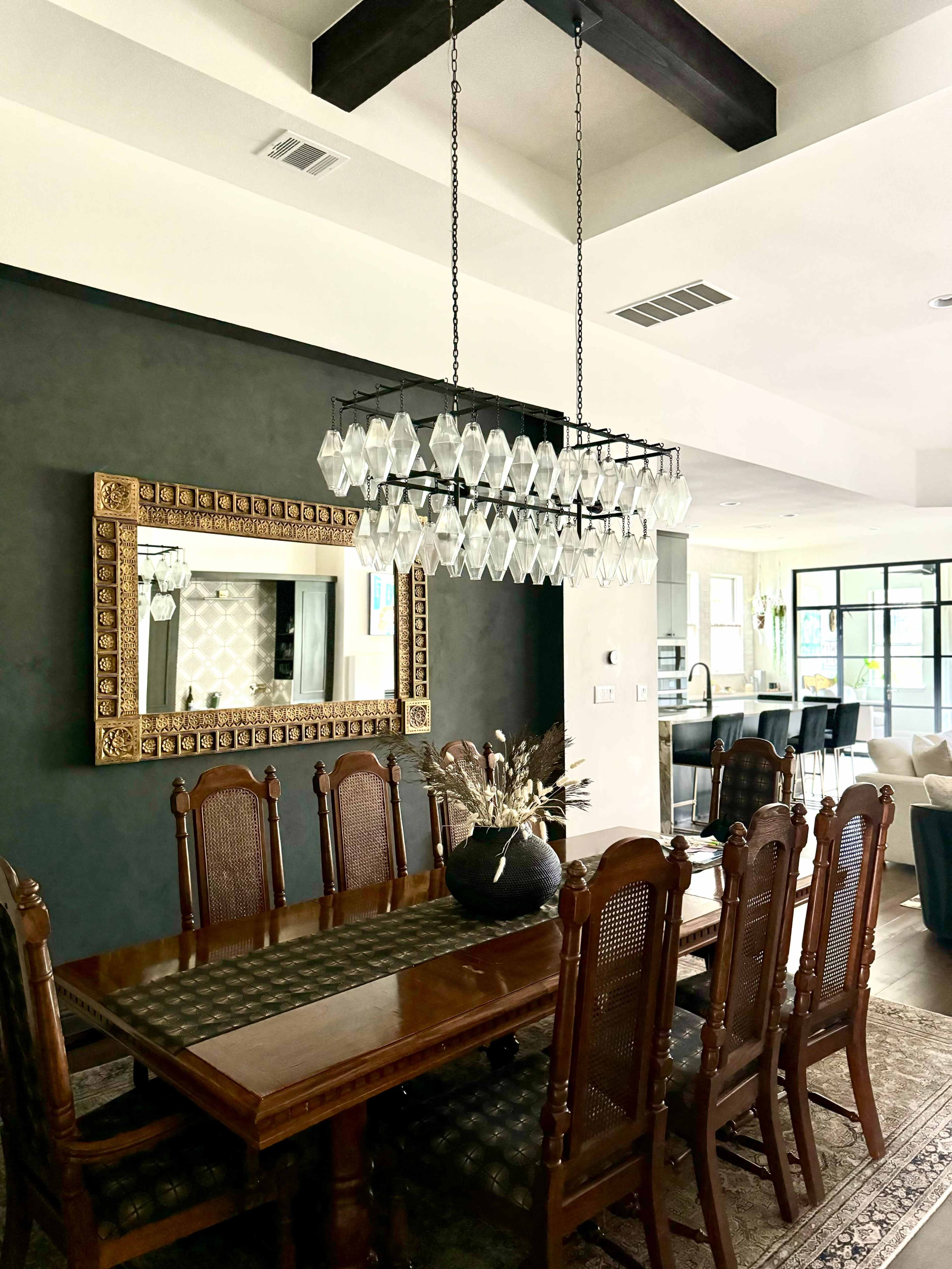 The image shows a dining room featuring a large wooden table surrounded by eight chairs, a decorative mirror on the wall, and a modern chandelier overhead.