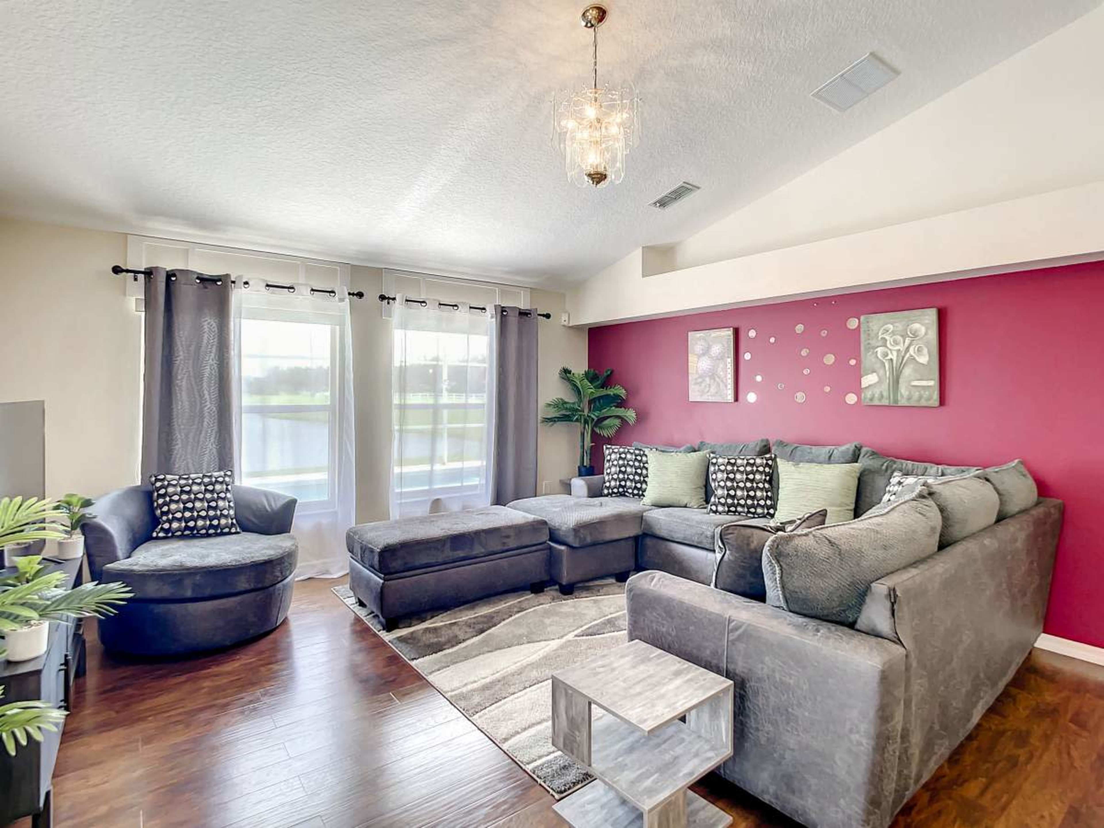 The room features a sectional sofa, an armchair, and large windows with sheer curtains, all arranged around a decorative wall in a blend of colors.