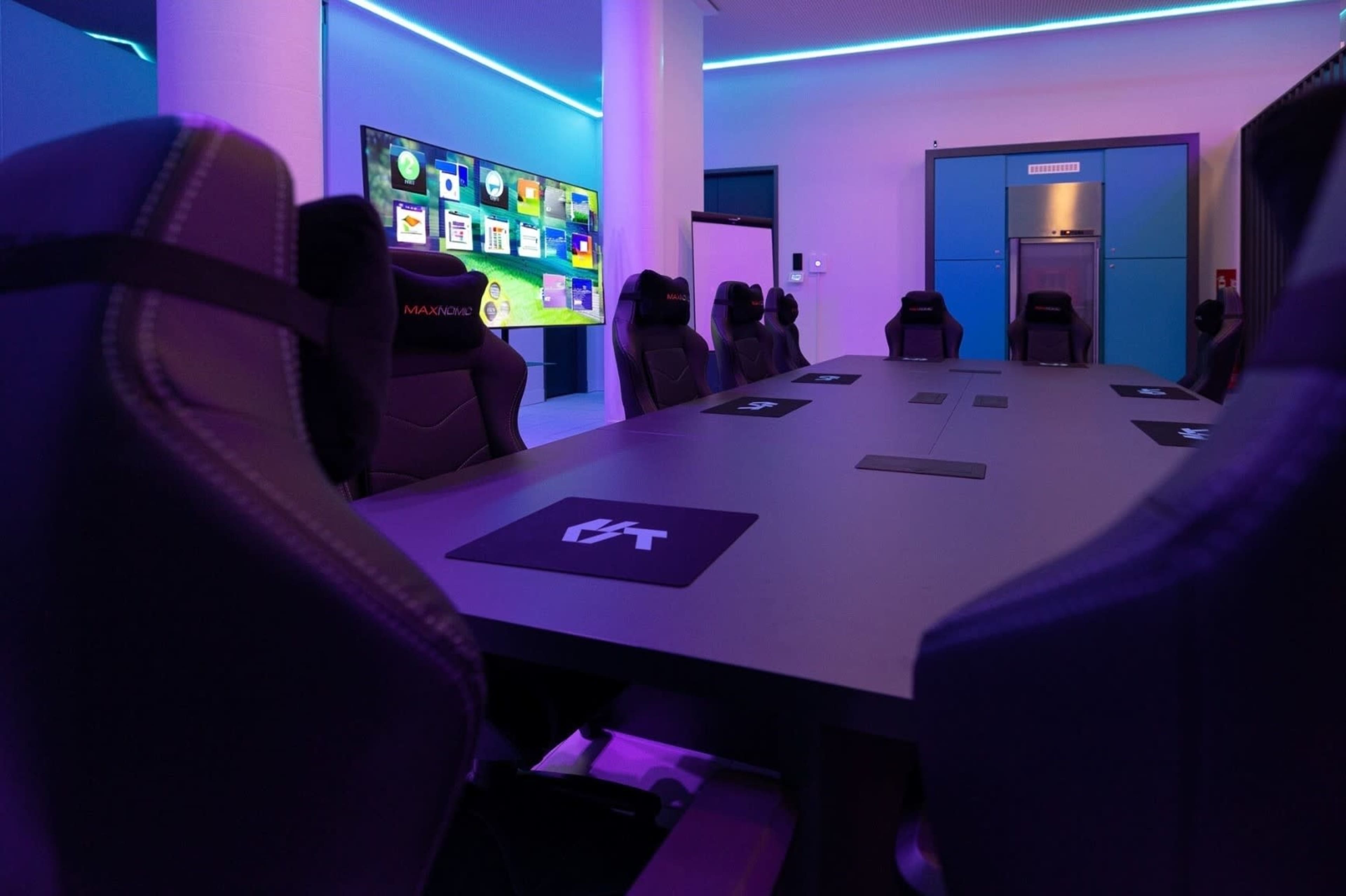A modern conference room features a long table surrounded by ergonomic chairs, with a large screen displaying colorful graphics on the wall.