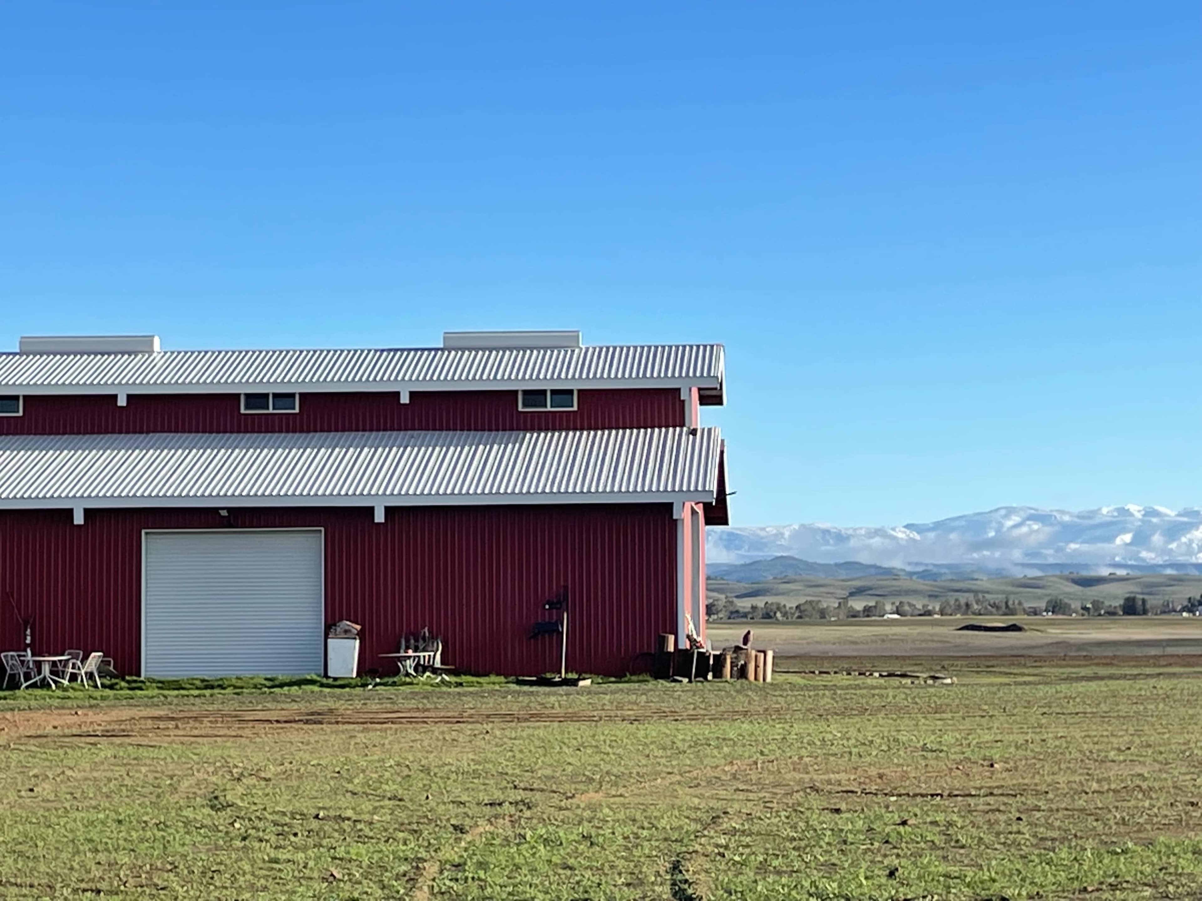 A red barn with a metal roof sits in a grassy field, with mountains visible in the background under a clear blue sky.
