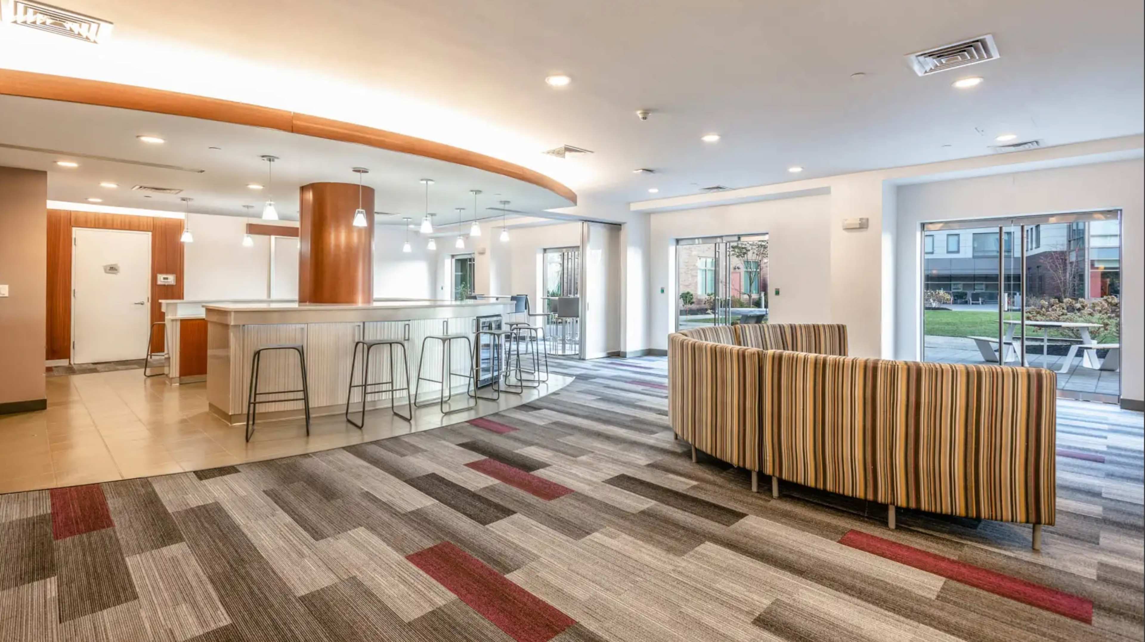 A modern common area with a striped sofa, a bar-style counter, and a dining area with high stools, all situated on patterned carpet.