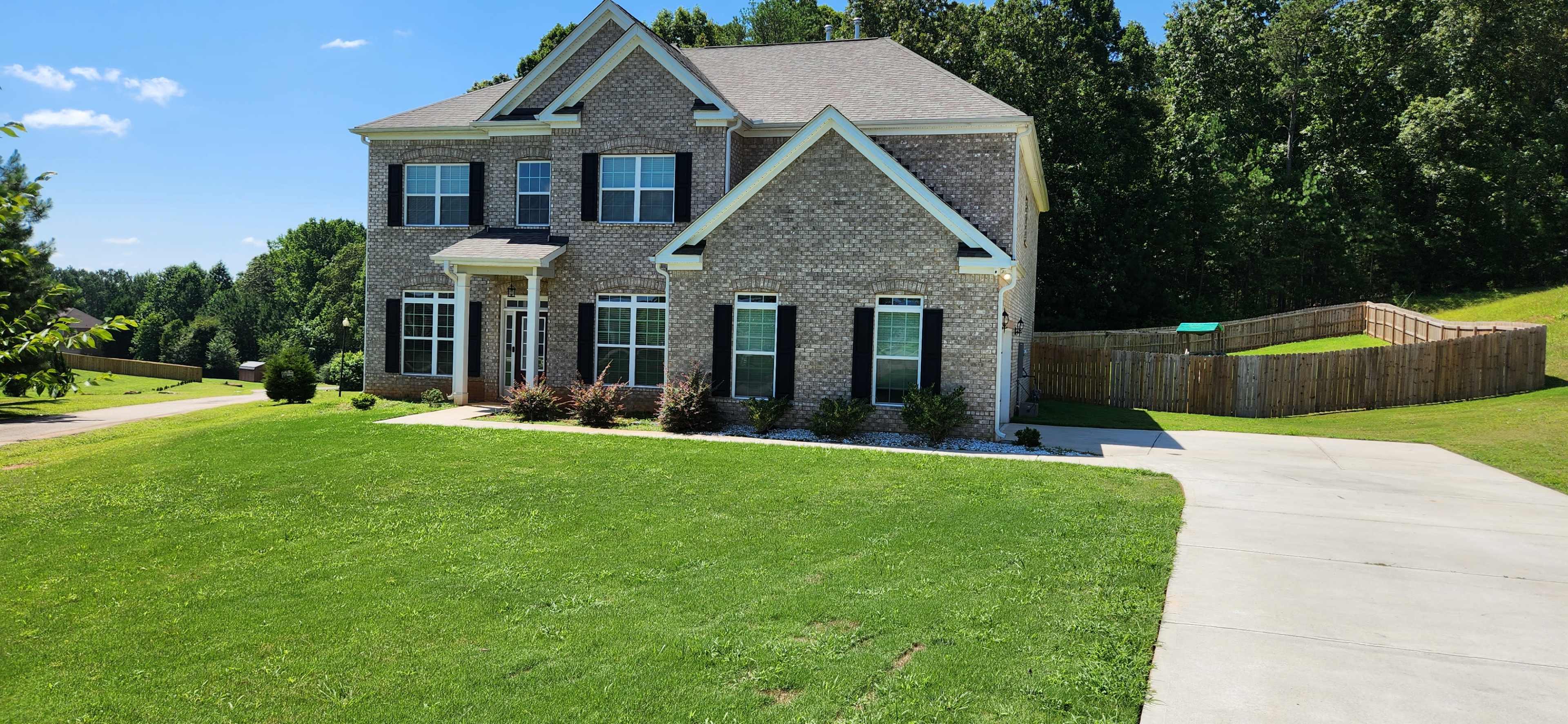 A two-story brick house with a neatly manicured lawn and a paved driveway is situated in a residential area.