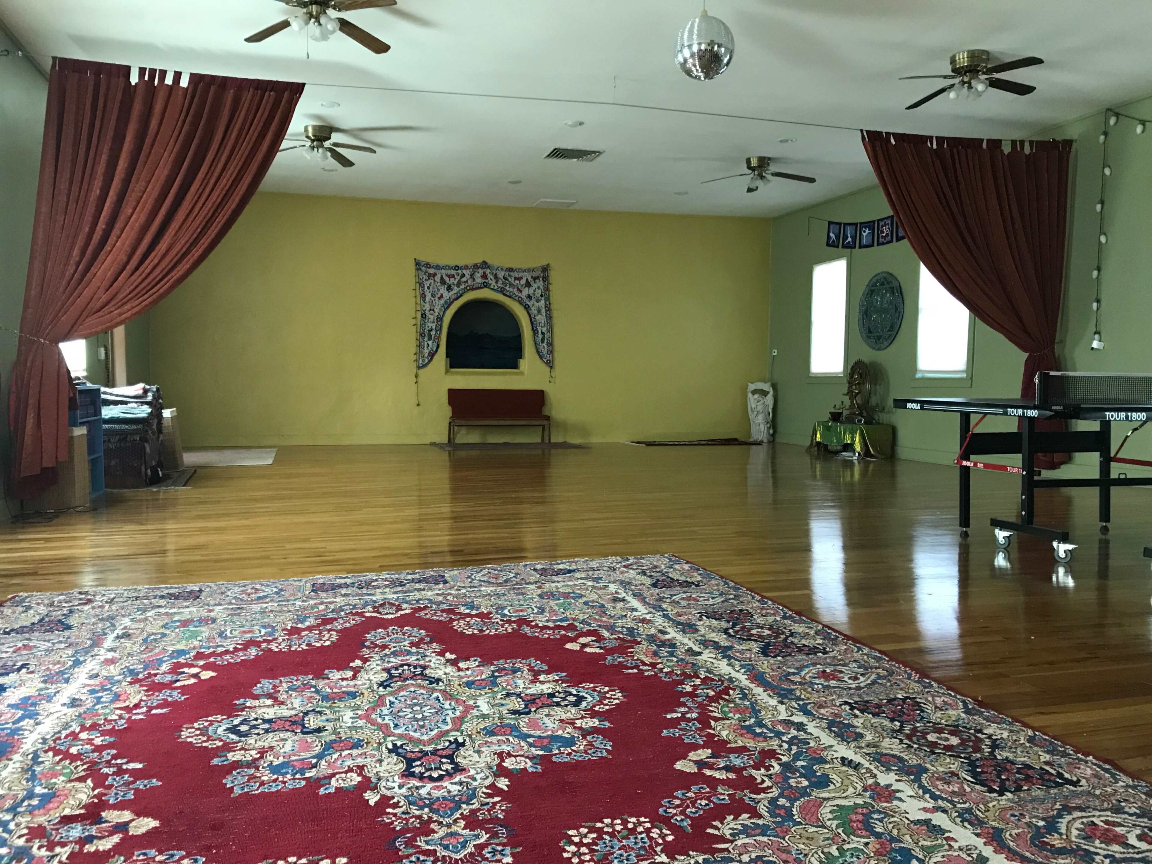 A spacious room with wooden flooring, adorned with a large red patterned rug, draped curtains, and a couple of ceiling fans.