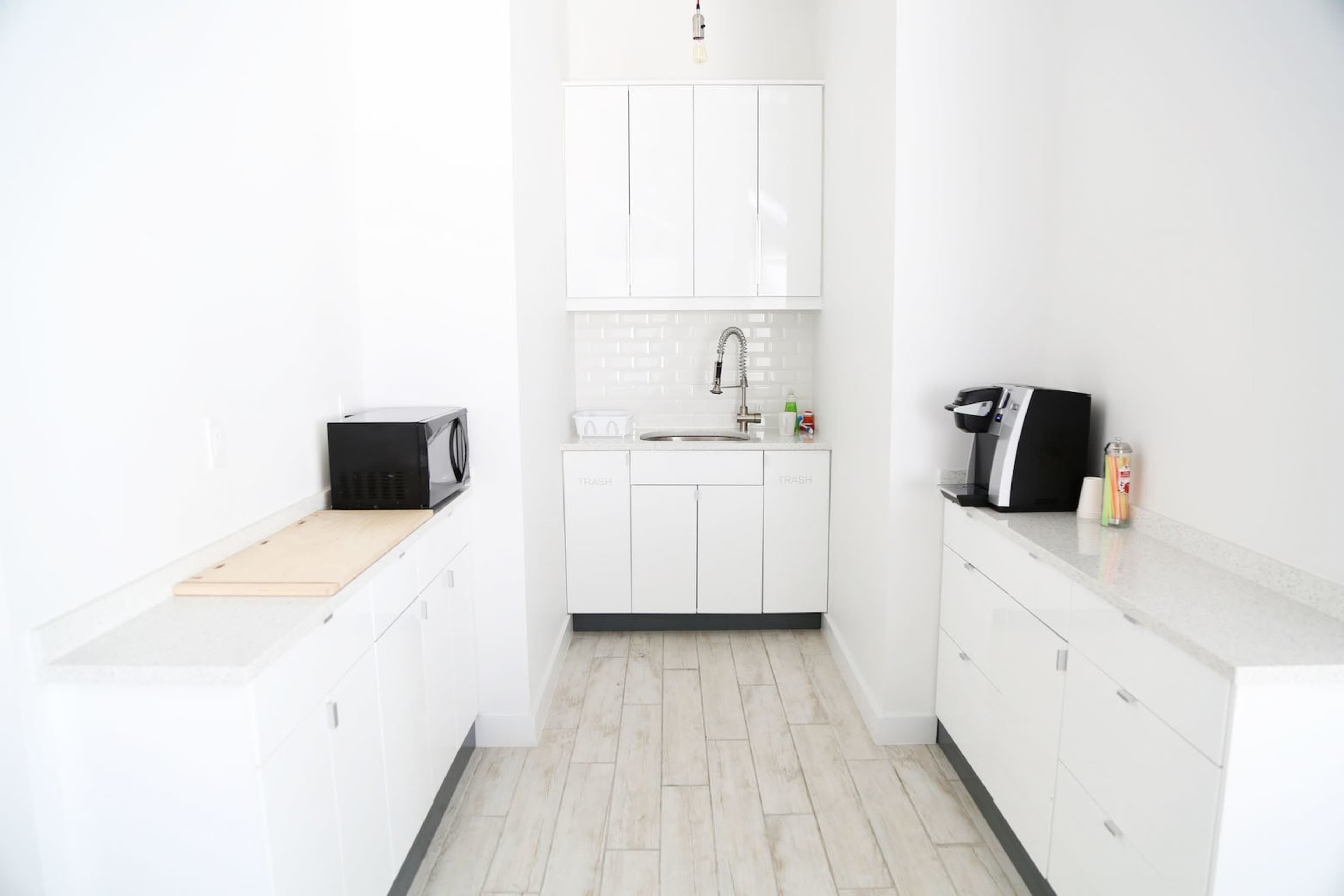 The image shows a modern kitchen area featuring white cabinets, a sink, a microwave, and a coffee machine with a light-colored tiled backsplash.