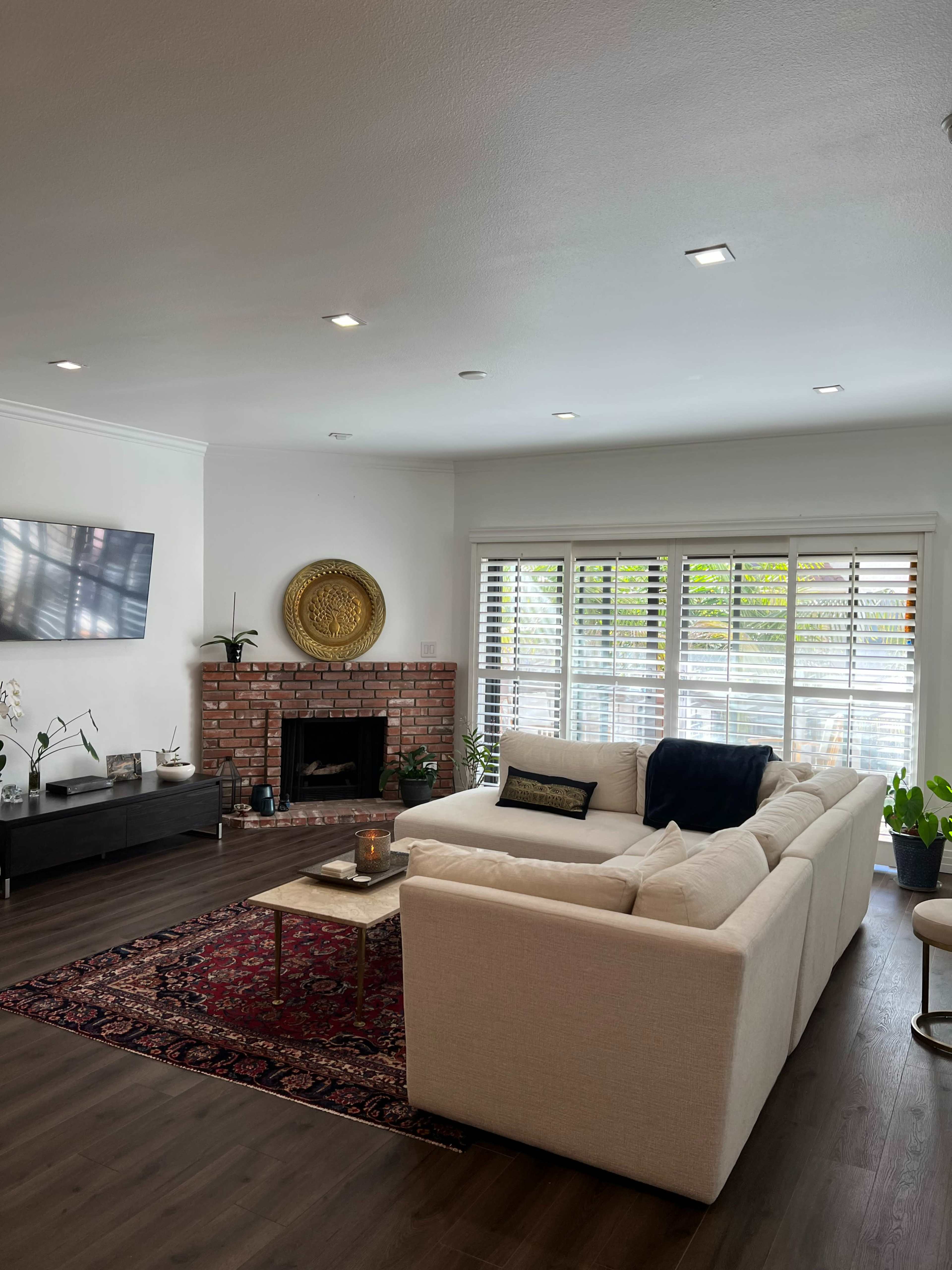 The living room features a sectional sofa facing a brick fireplace, with large windows adorned with blinds and a decorative wall piece.