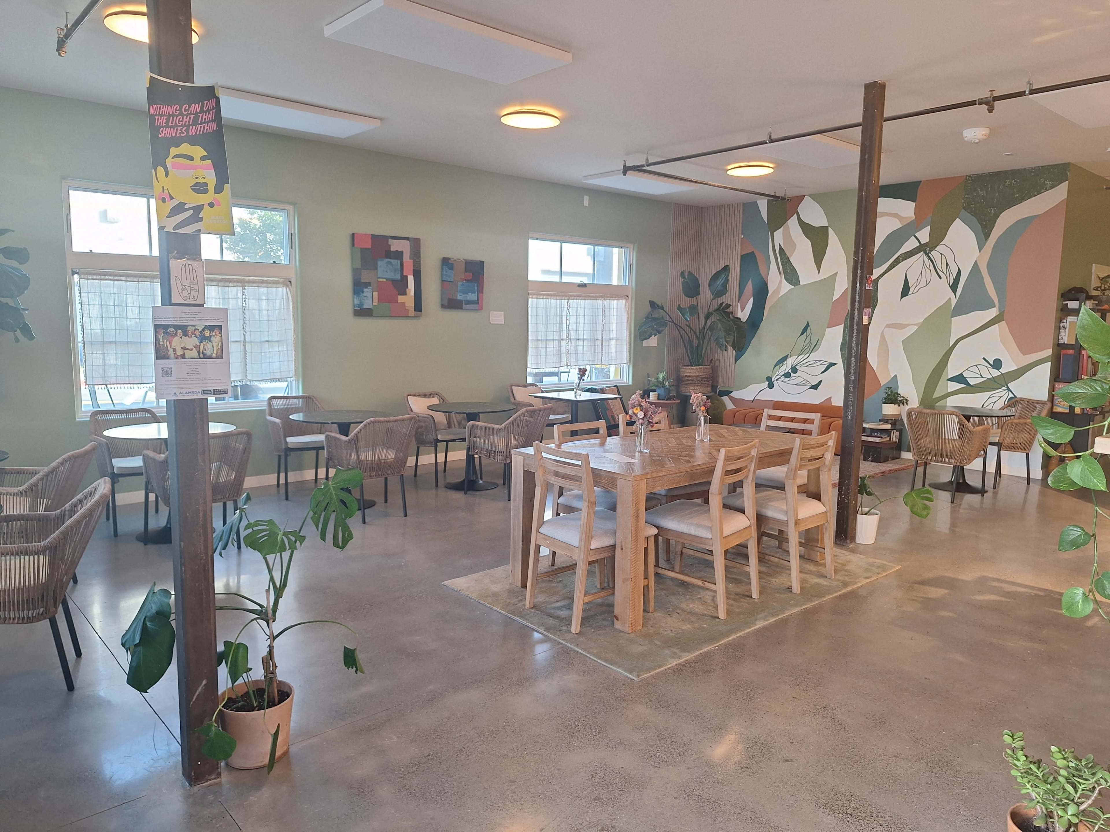 The image shows a spacious café with wooden tables surrounded by plants and a colorful mural on the wall.