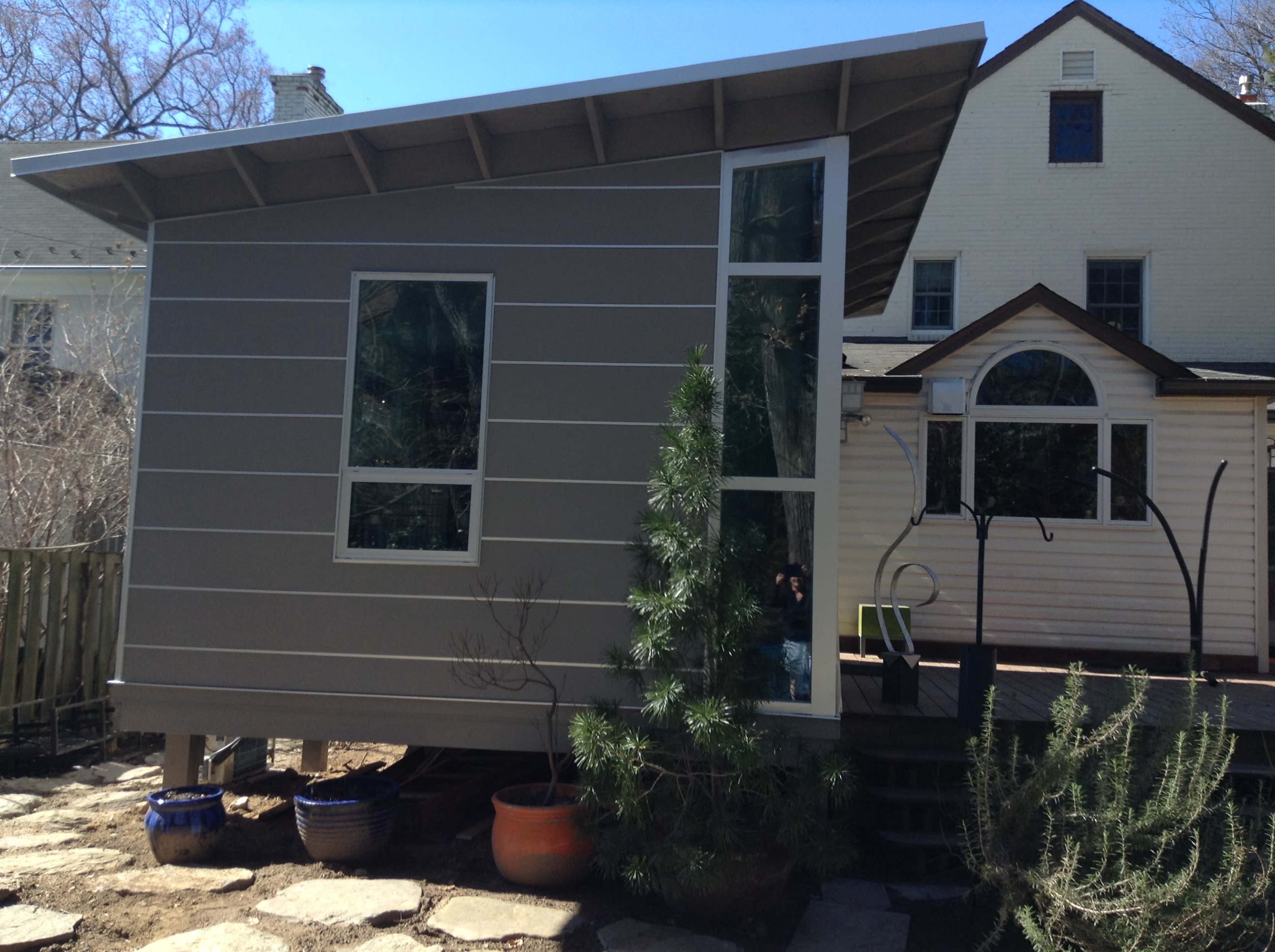 A modern gray shed with large windows situated next to a traditional white house, surrounded by landscaped stone pathways and plants.
